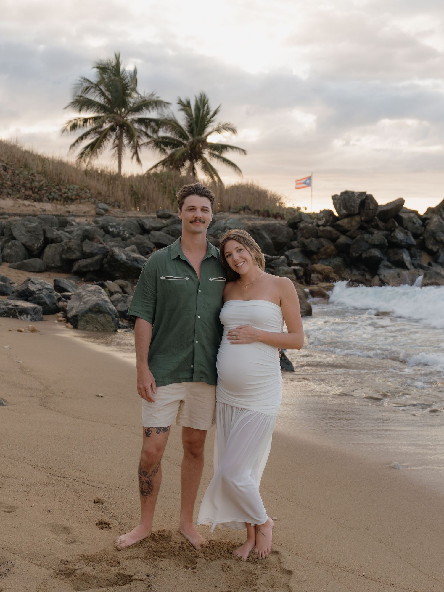 Couple poses on tropical beach at sunset, woman in white maternity dress, man in green shirt, palm trees and rocks behind them.