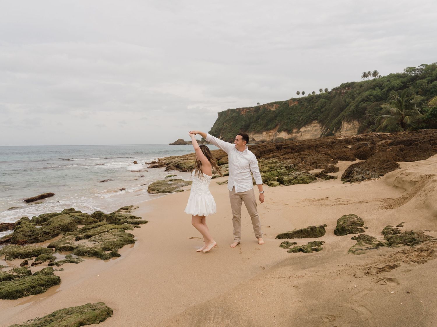 Couple dancing on a sandy beach with rocky cliffs and ocean waves in the background on a cloudy day.
