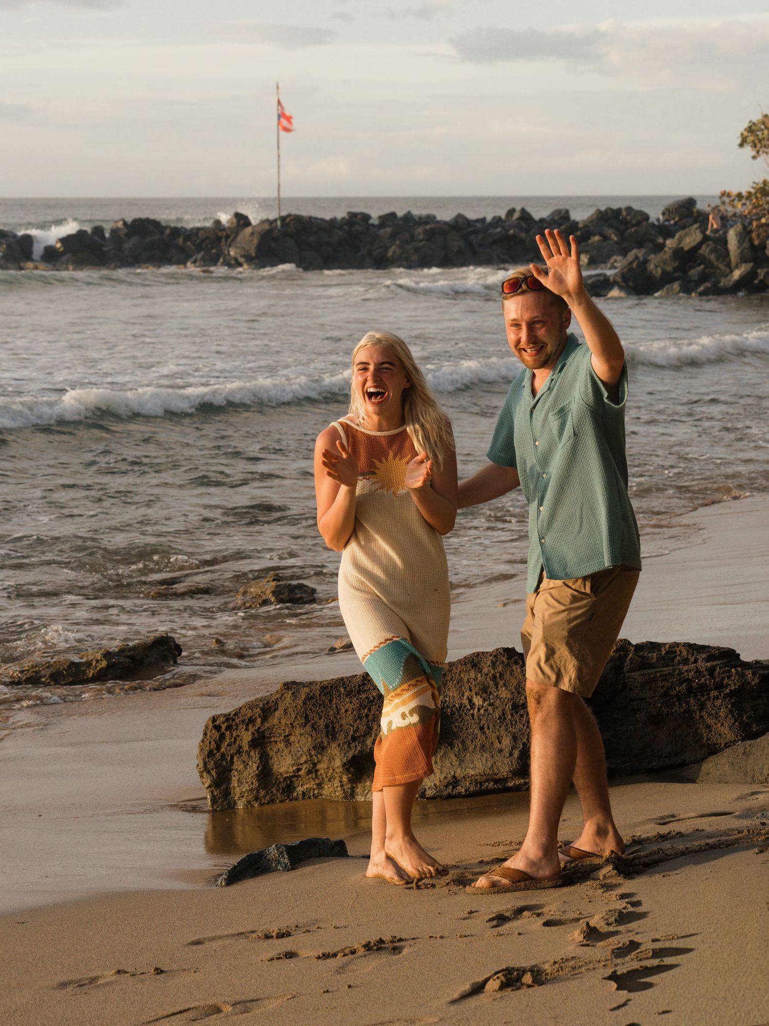A joyful couple waving and laughing on a sandy beach at sunset with ocean waves and rocks behind them.