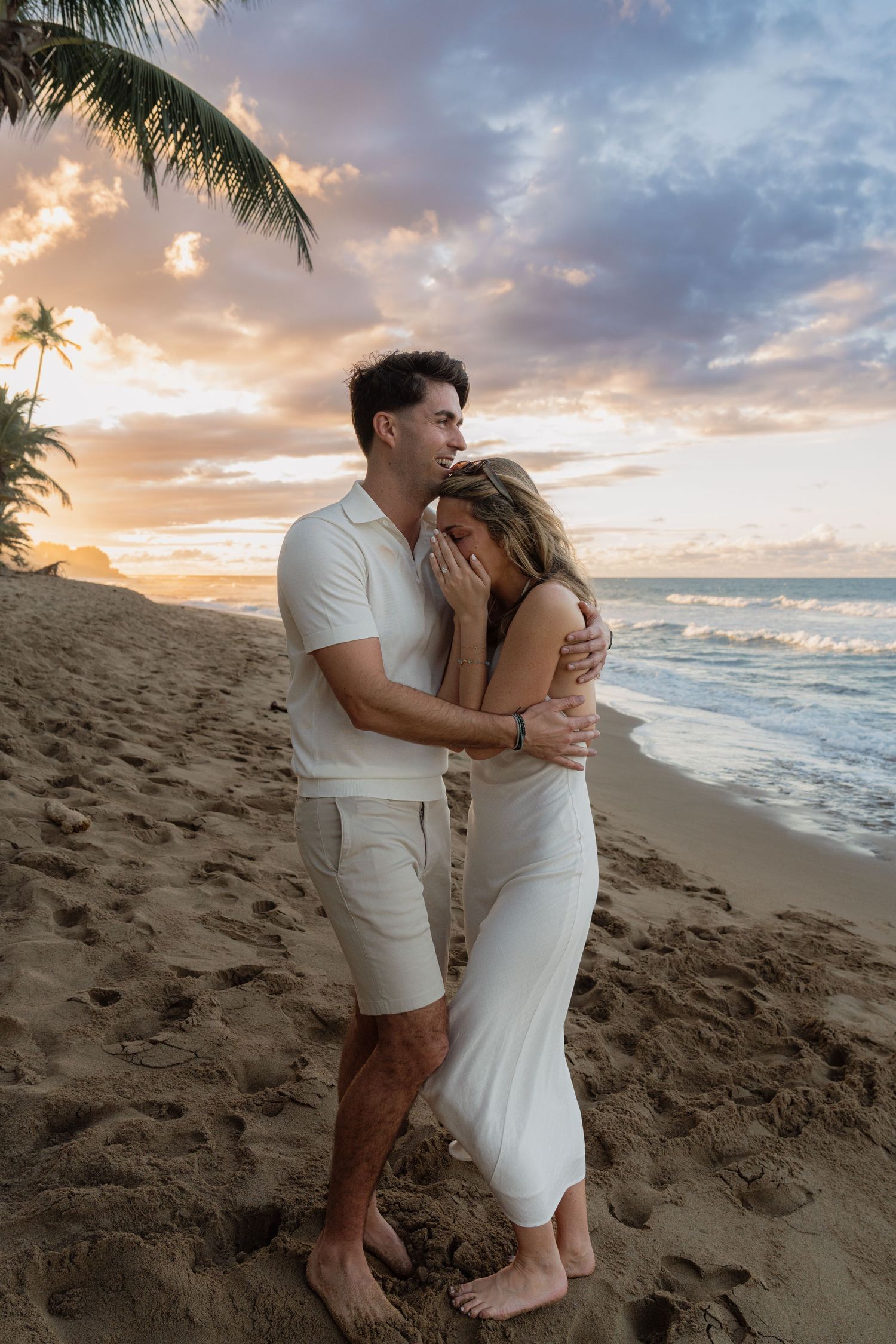 Couple embracing on a tropical beach at sunset, wearing white outfits, with palm trees and colorful sky.