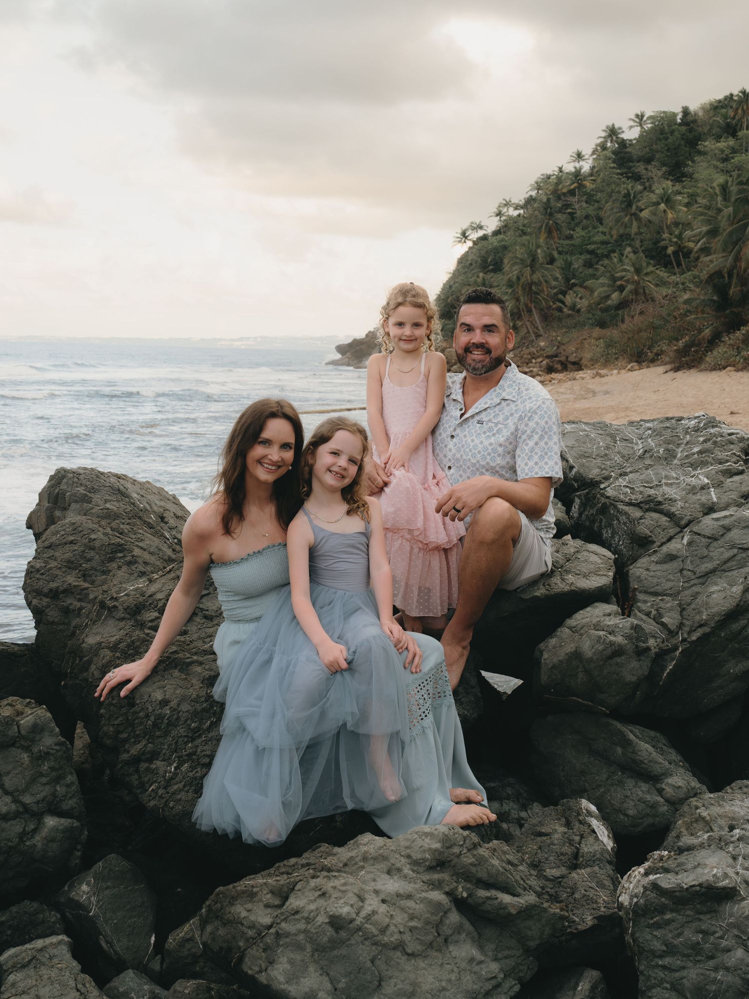 Family of four posing on rocky beach with ocean waves and lush green hillside in background.