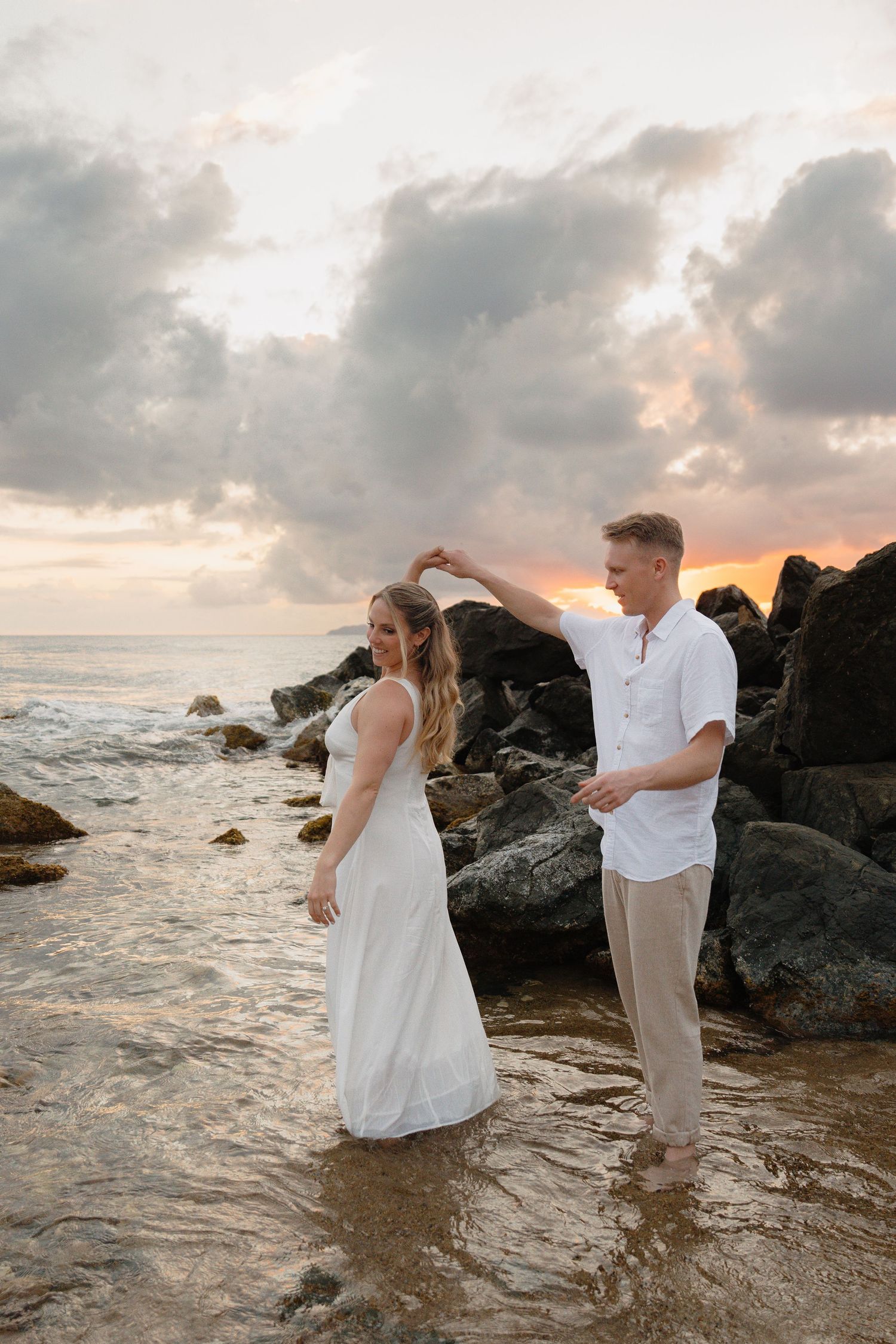 Couple dancing on a rocky beach at sunset, woman in white dress, man in white shirt, waves and dramatic cloudy sky.