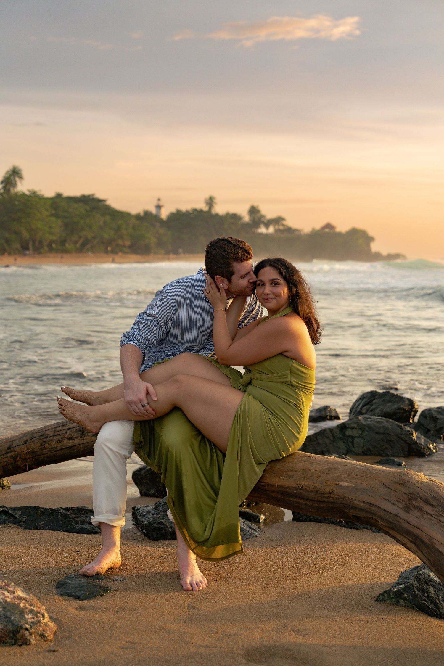 Couple sharing a romantic sunset moment on driftwood at a tropical beach, woman in green dress smiling.