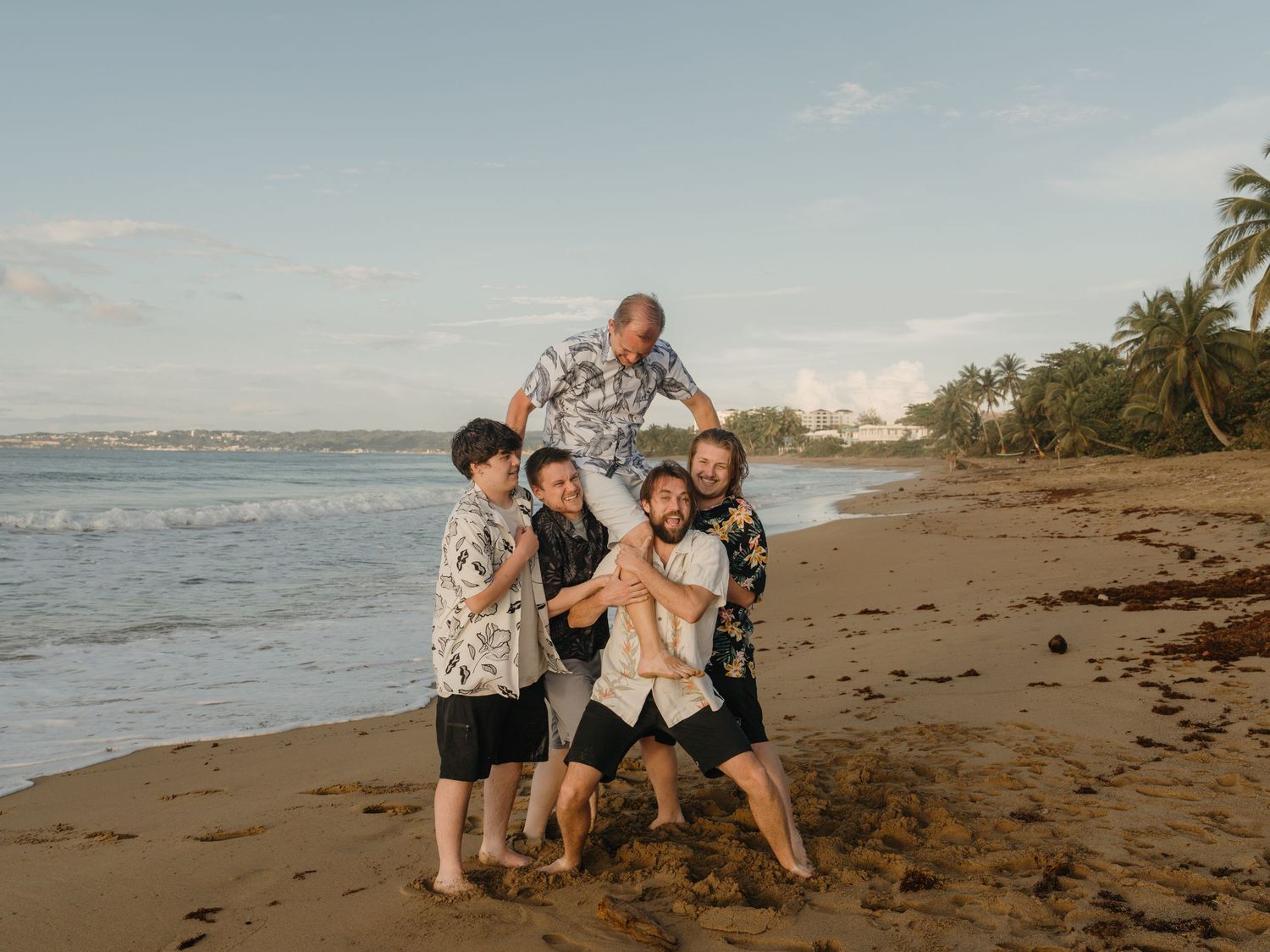 Five men pose playfully on a tropical beach at sunset, wearing Hawaiian shirts and laughing together.