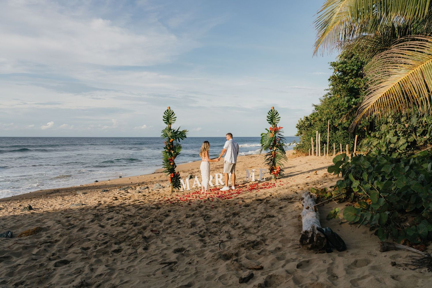 Couple standing at a romantic beach wedding setup with floral arches and MARRY ME sign on tropical sandy shore.