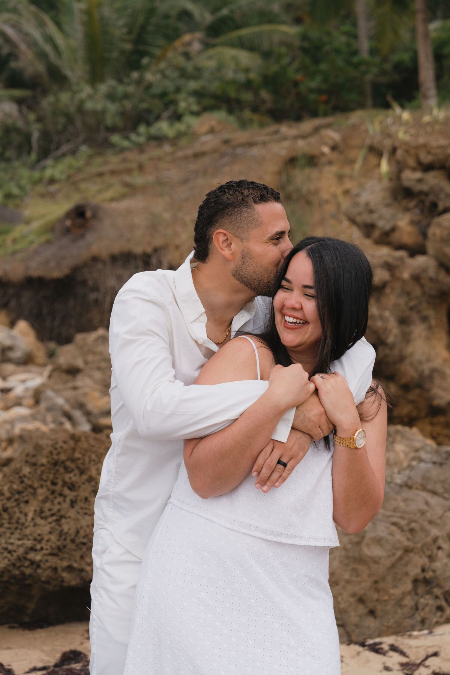 Couple in white outfits embracing on a rocky beach, man kissing woman's cheek as she laughs joyfully.
