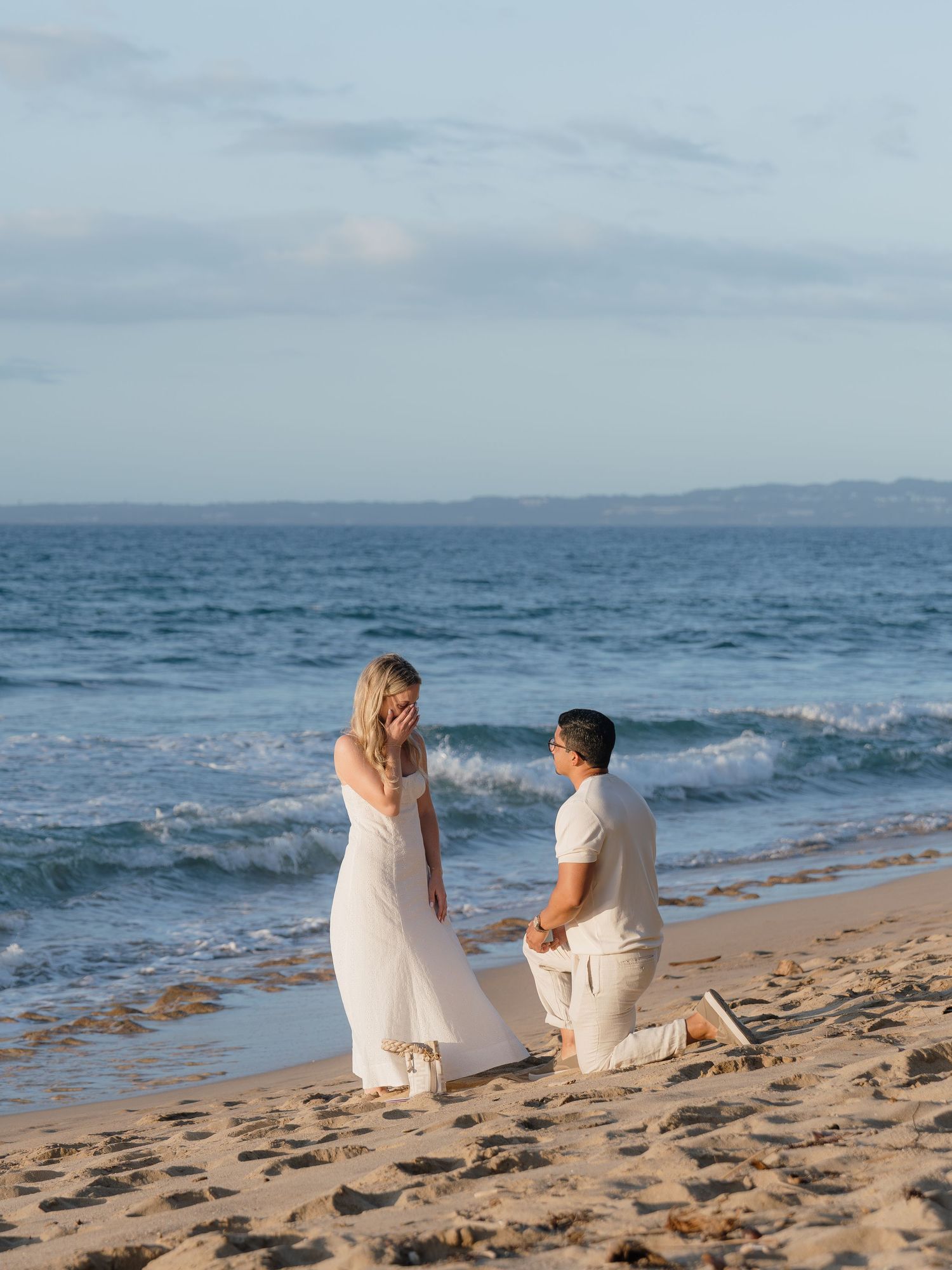 Man proposes on bended knee to woman in white dress on sandy beach at sunset with ocean waves behind them.