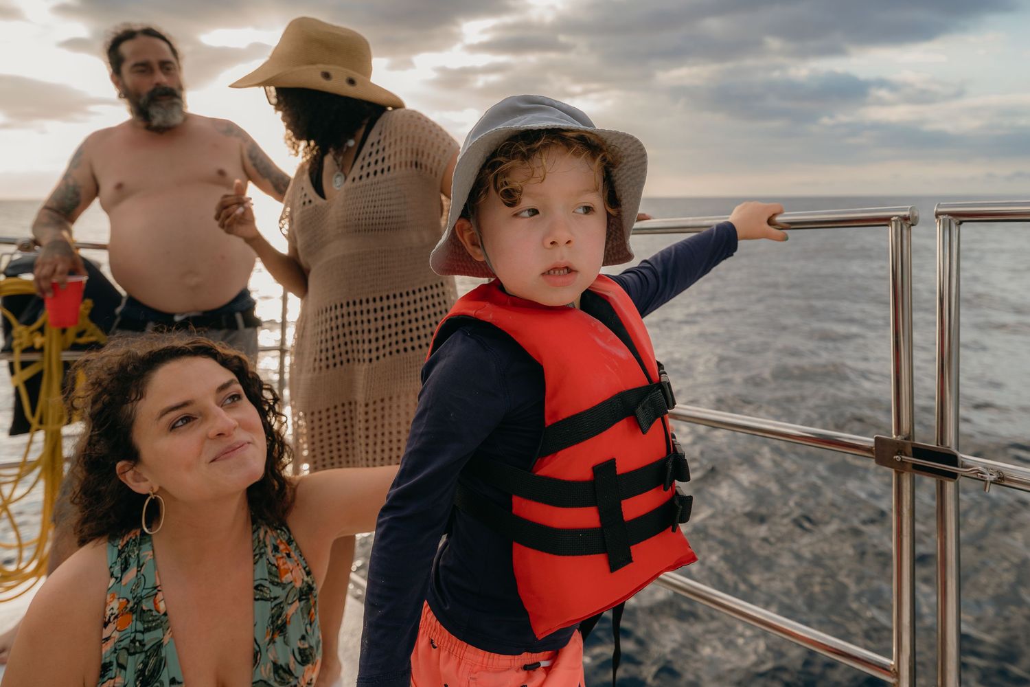 A child in a life jacket points from a boat deck while adults stand nearby against a cloudy ocean backdrop.