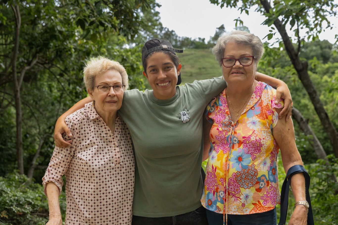 Three women smiling outdoors in a lush green tropical setting, posing together with arms around each other.
