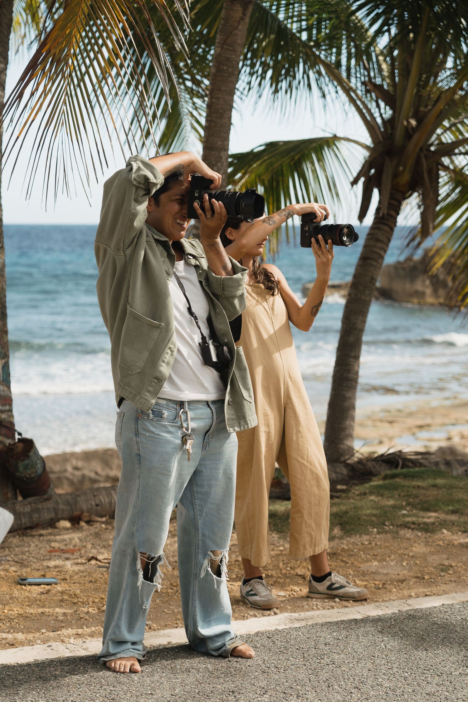 Two photographers capturing shots near palm trees on a tropical beach with turquoise ocean waves in the background.