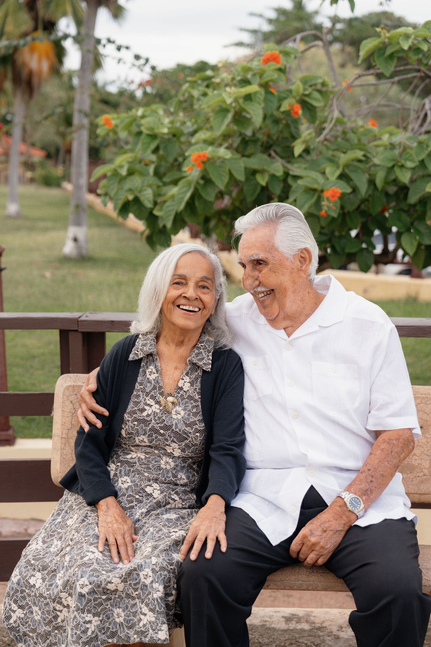 Happy elderly couple sitting on a bench in a lush garden, smiling and embracing on a sunny day.