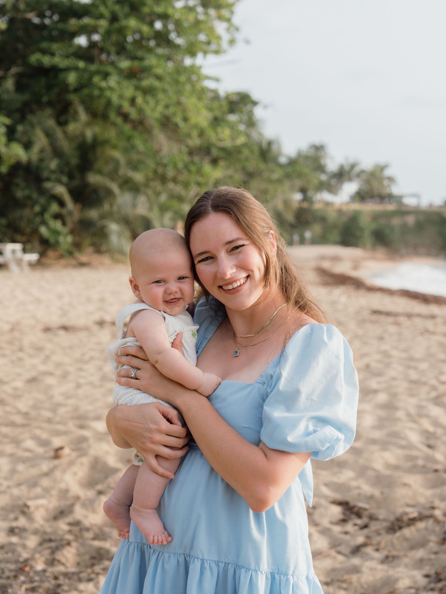 A smiling young woman in a blue dress holds a happy baby on a sandy beach with lush green trees behind them.