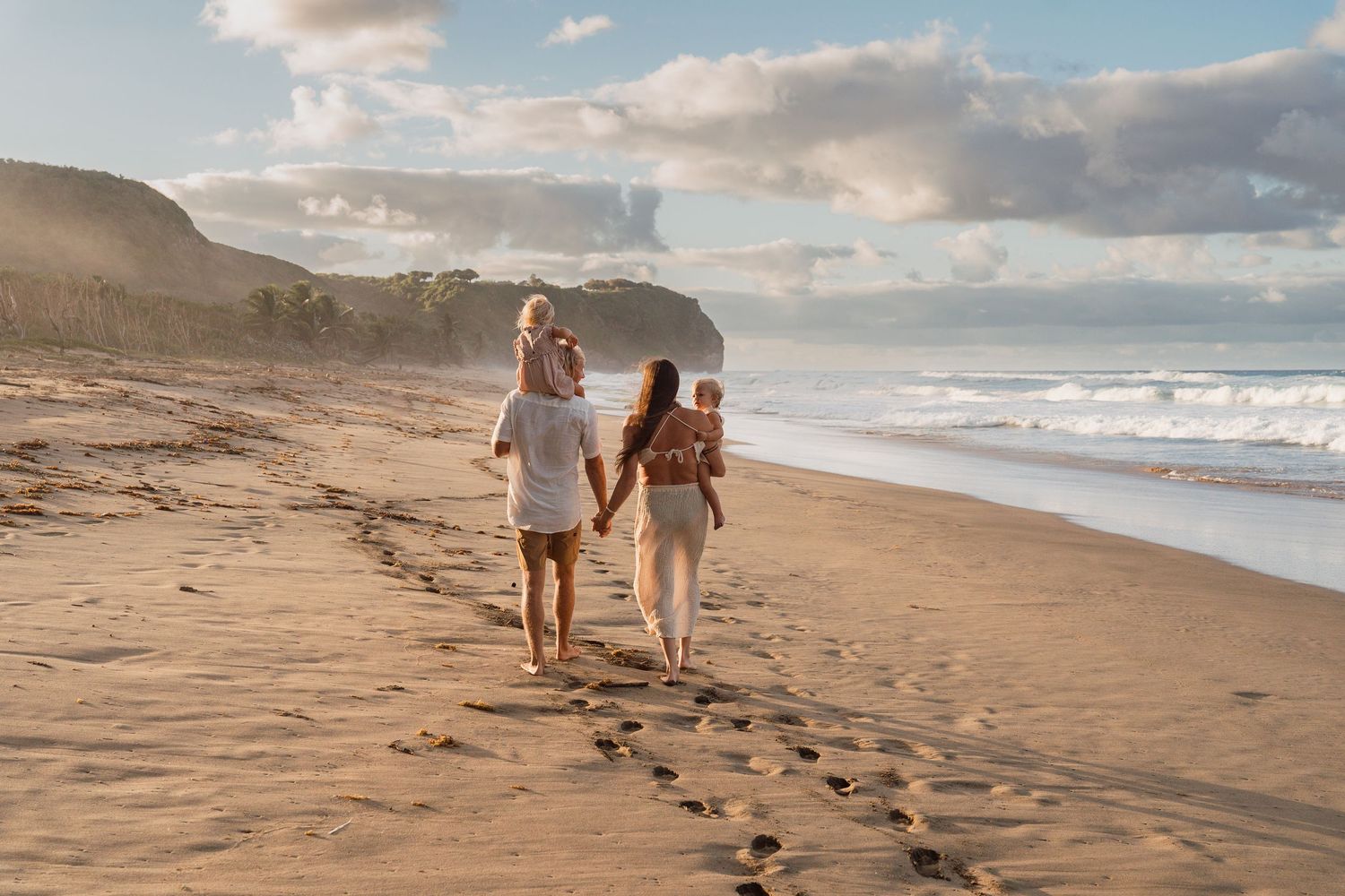 Two women walk along a sandy beach at sunset, one carrying a child on her hip and another on her shoulders.