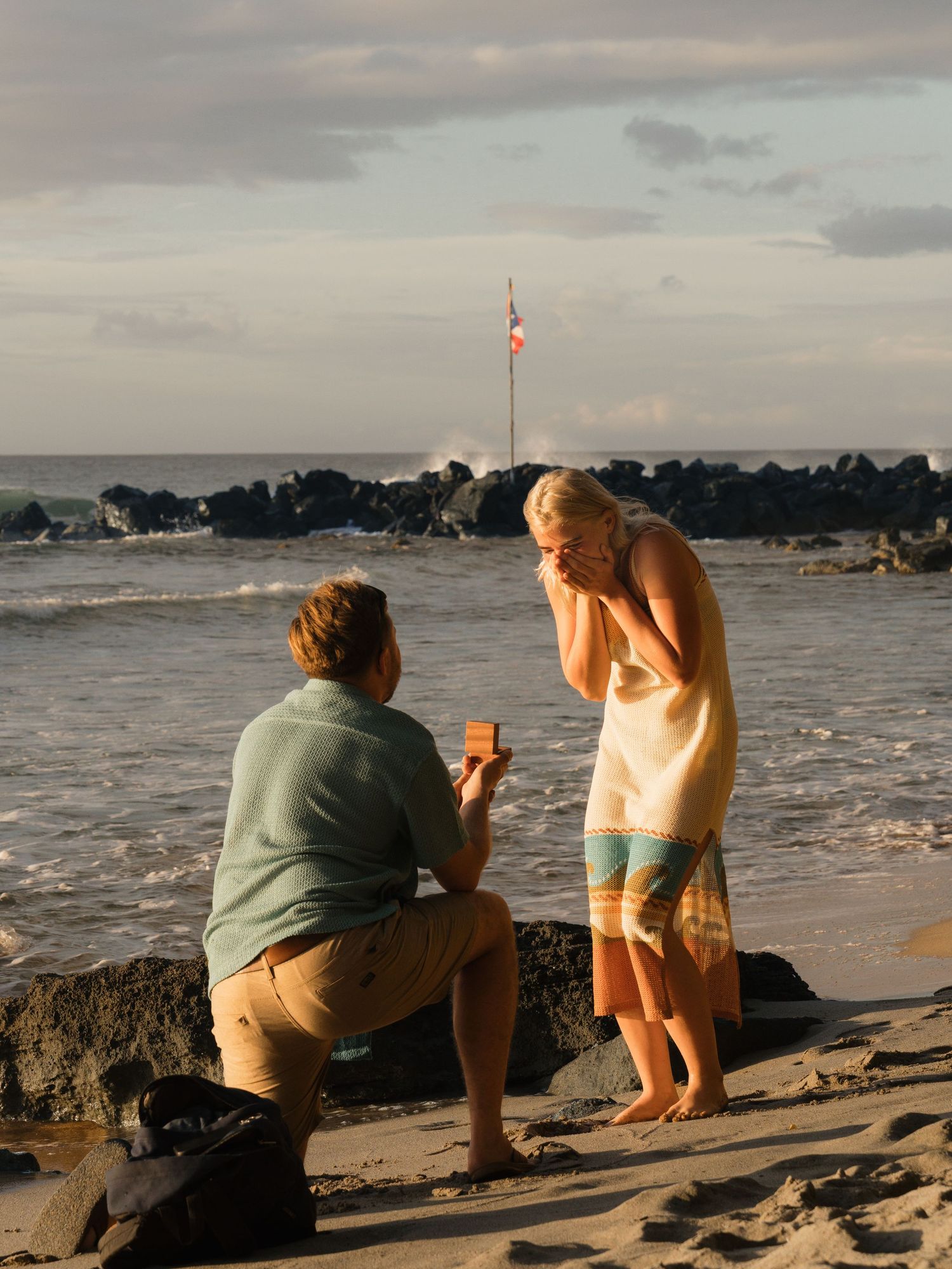 Man proposes to woman on a rocky beach at sunset, holding a ring box while kneeling on one knee.