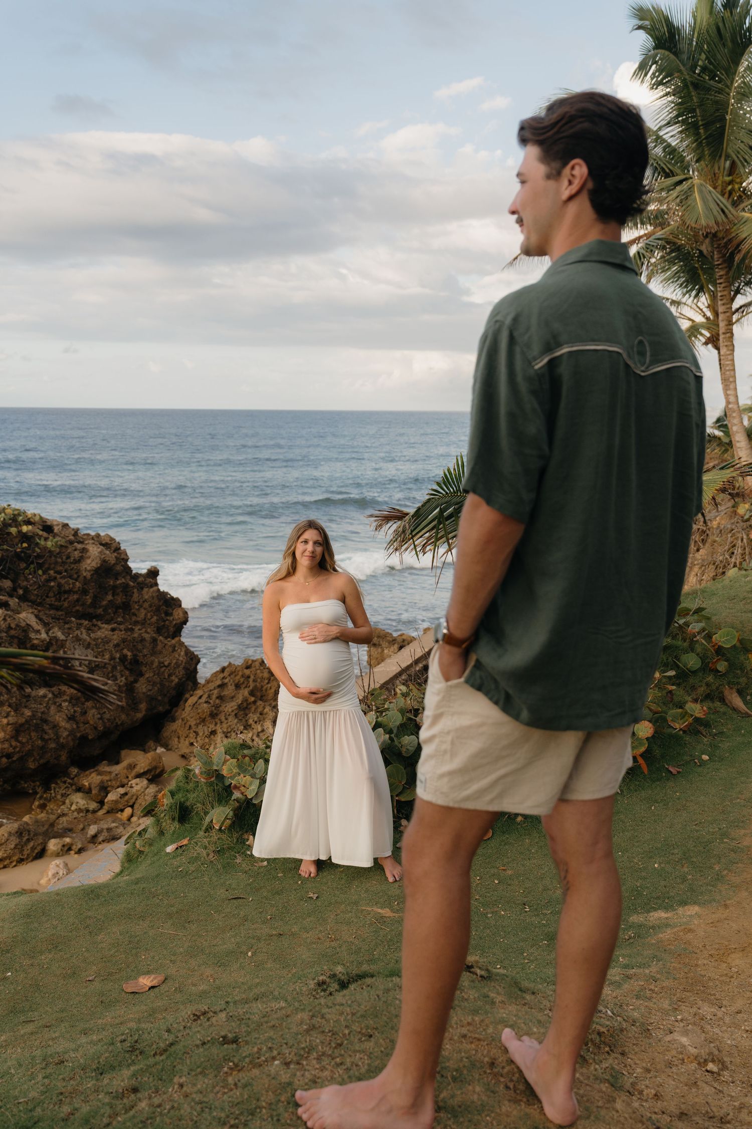 Pregnant woman in white dress poses near ocean cliffs while man in green shirt watches during maternity photoshoot.