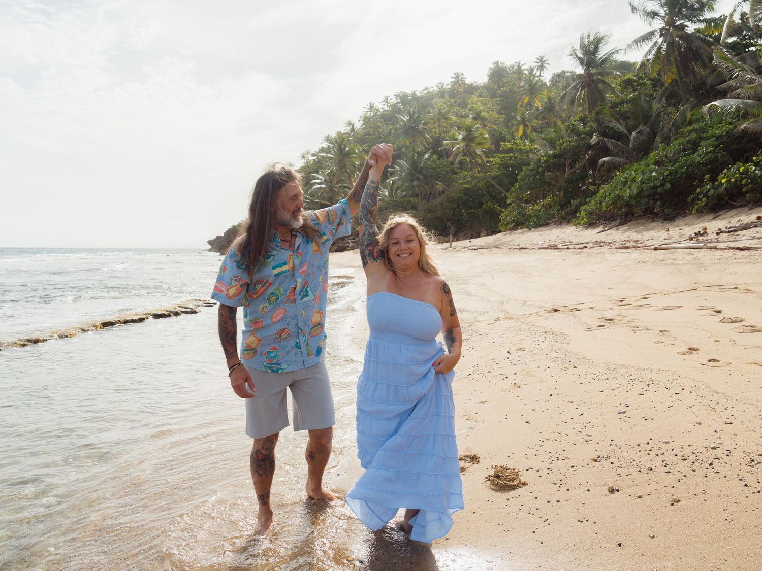 Couple celebrating on a tropical beach, man in blue Hawaiian shirt raises woman's hand, palm trees in background.