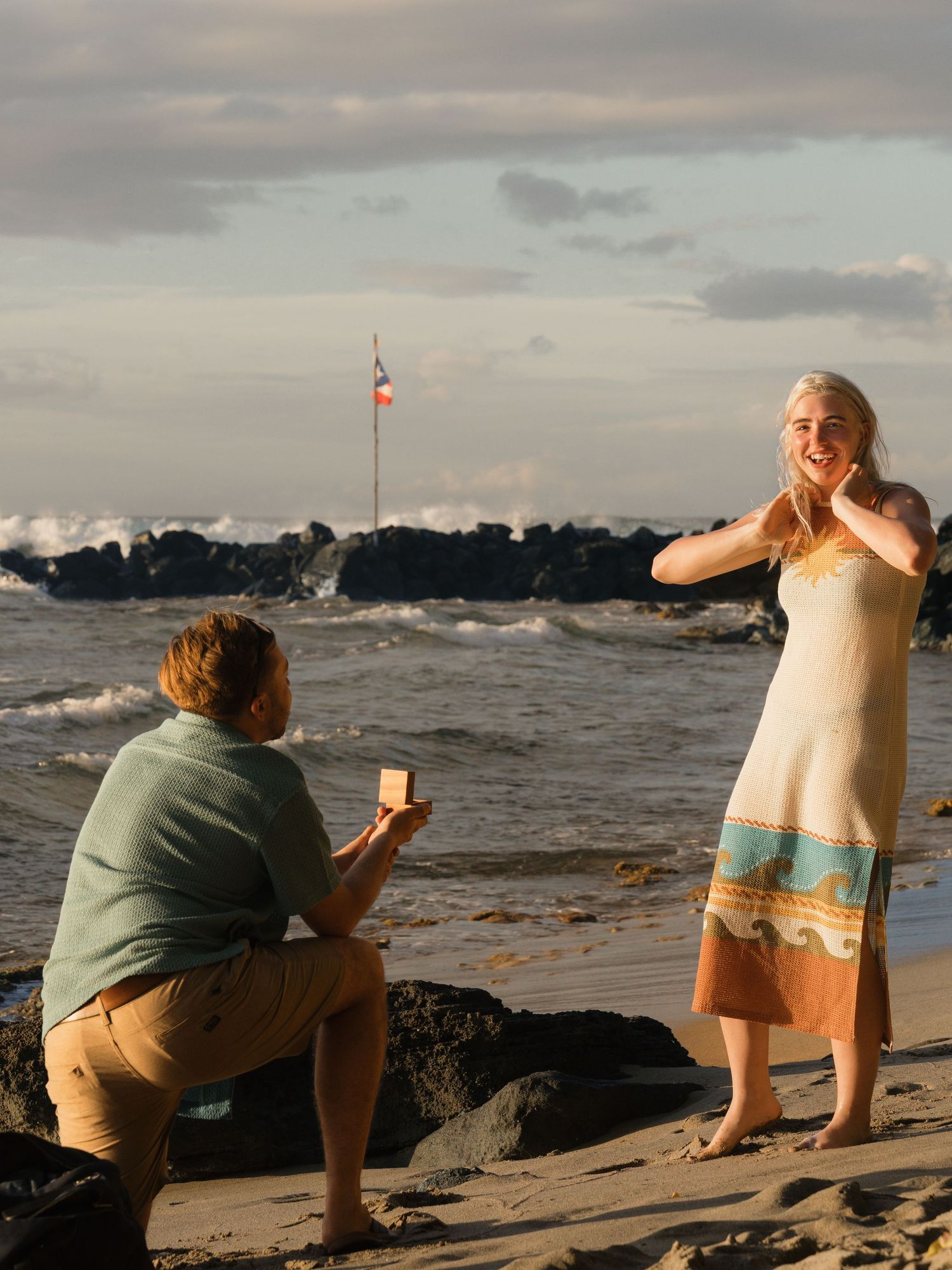 Man kneeling on beach proposing to woman in dress near rocky shore with flag and ocean waves behind them.