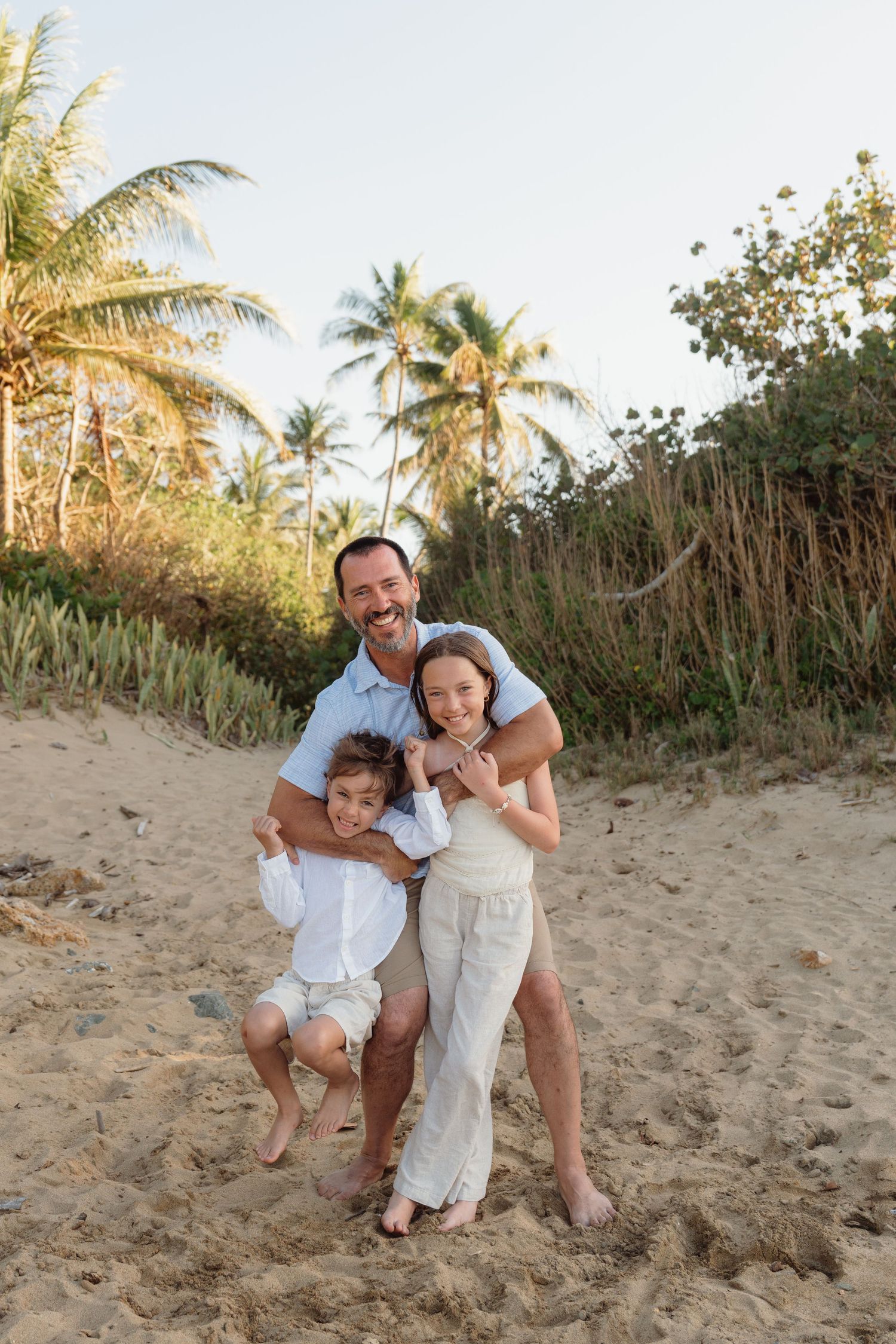 A smiling father hugs his two children on a sandy tropical beach surrounded by palm trees at golden hour.