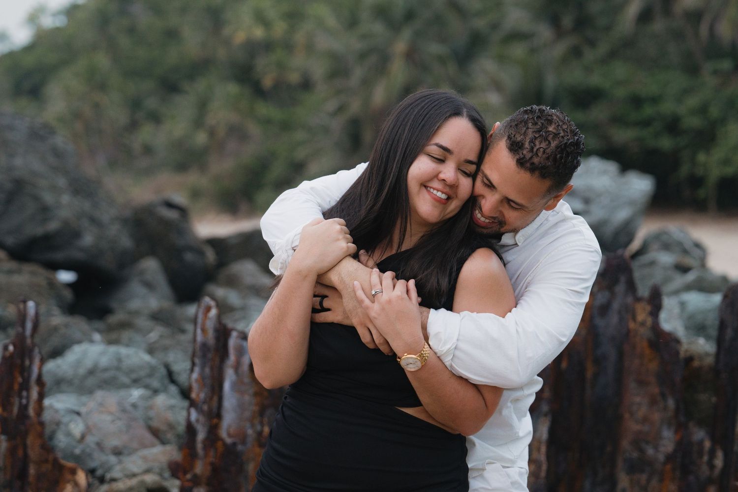 Couple embracing and smiling on a rocky beach, woman in black dress, man in white shirt.