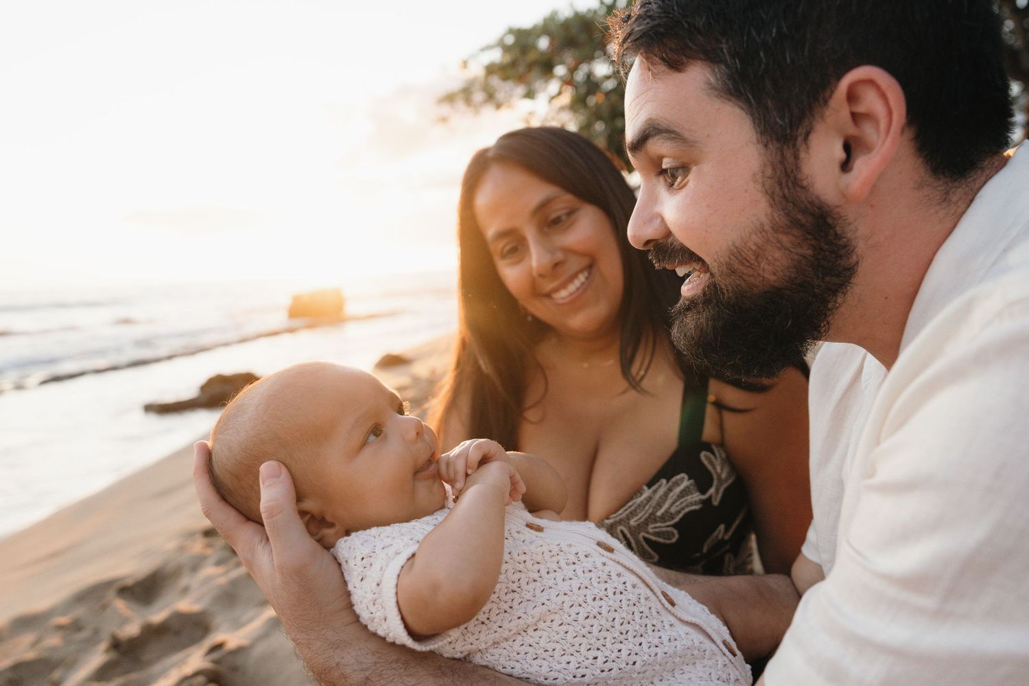 A couple smiles at their newborn baby on a sunny beach at golden hour.