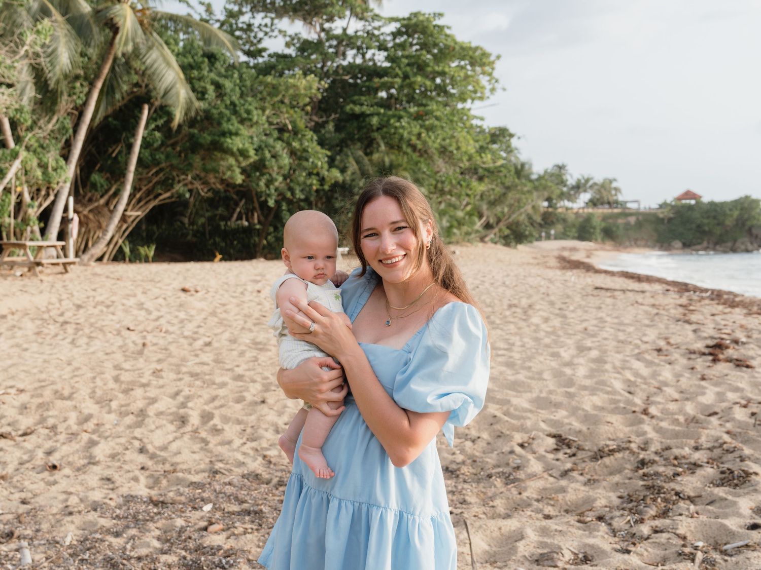 A smiling young woman in a blue dress holds a baby on a sandy tropical beach with lush green trees behind her.