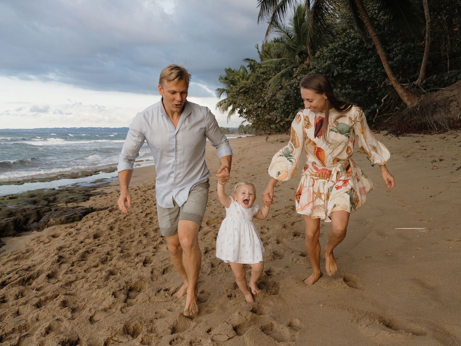 Family of three walking on tropical beach, parents swinging toddler in white dress between them near palm trees.