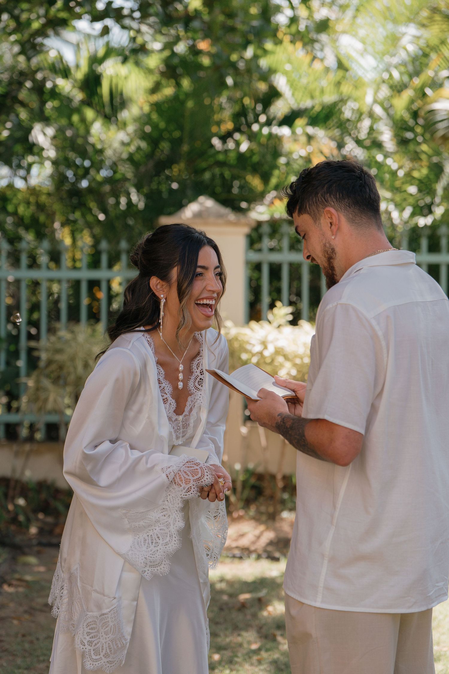 Bride in white lace gown laughs joyfully as groom reads wedding vows outdoors in tropical garden setting.