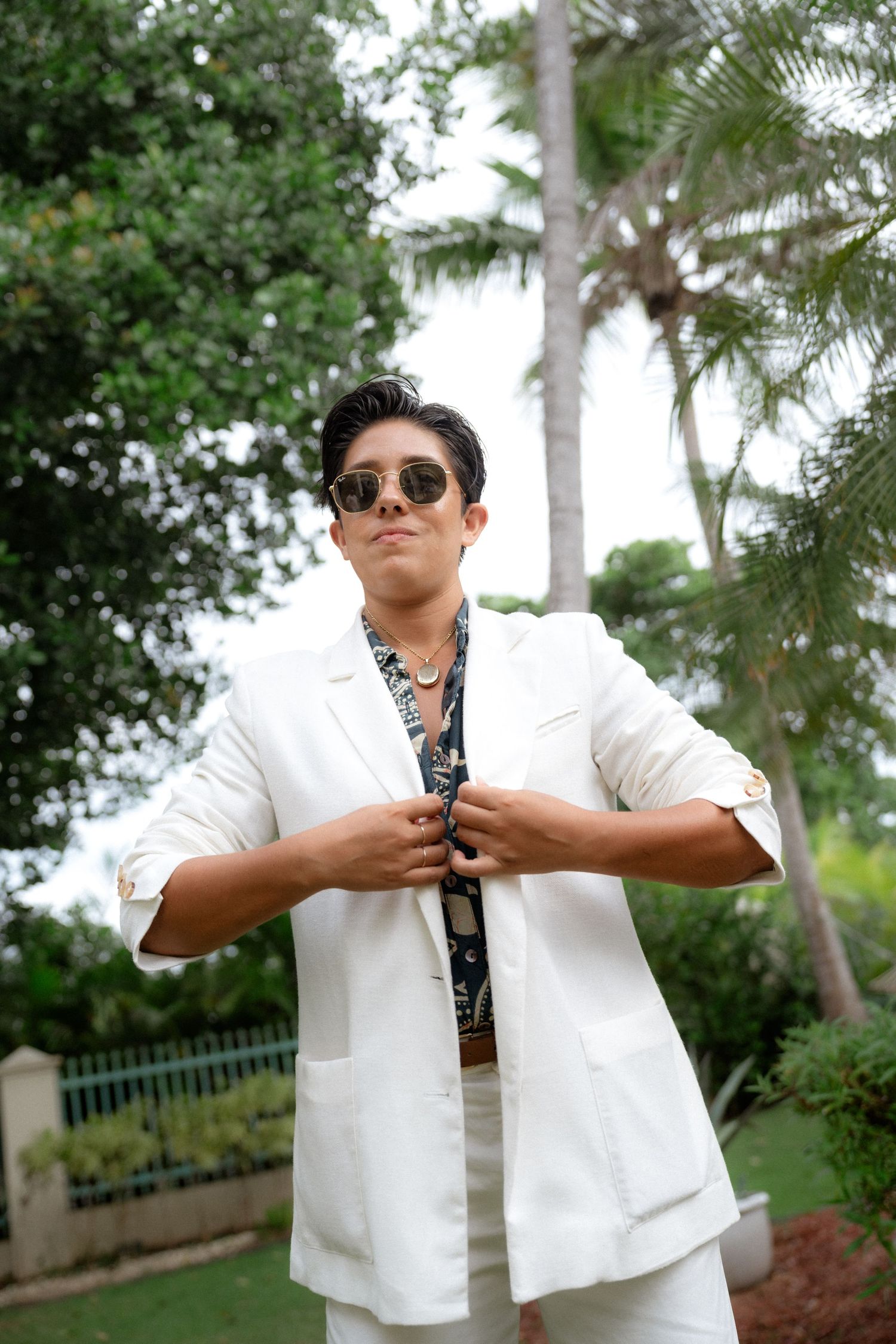 Stylish young man in white suit and sunglasses adjusting jacket outdoors with palm trees in background.
