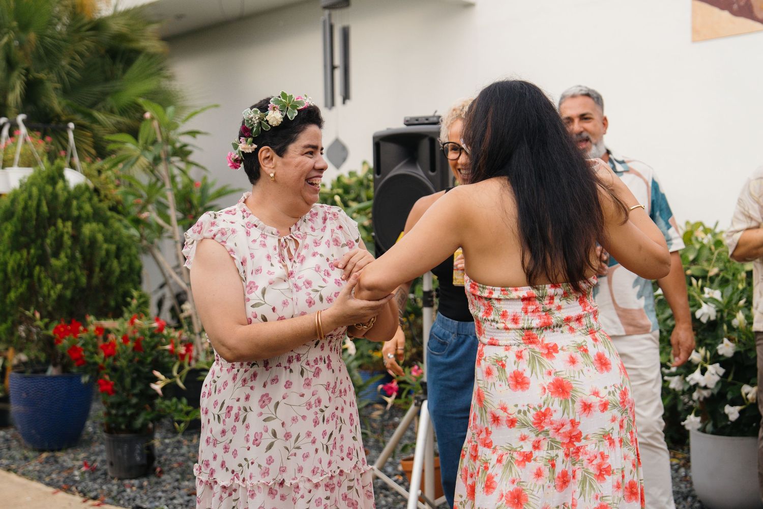 Two women in floral dresses dance joyfully outdoors at a garden party, one wearing a flower crown.