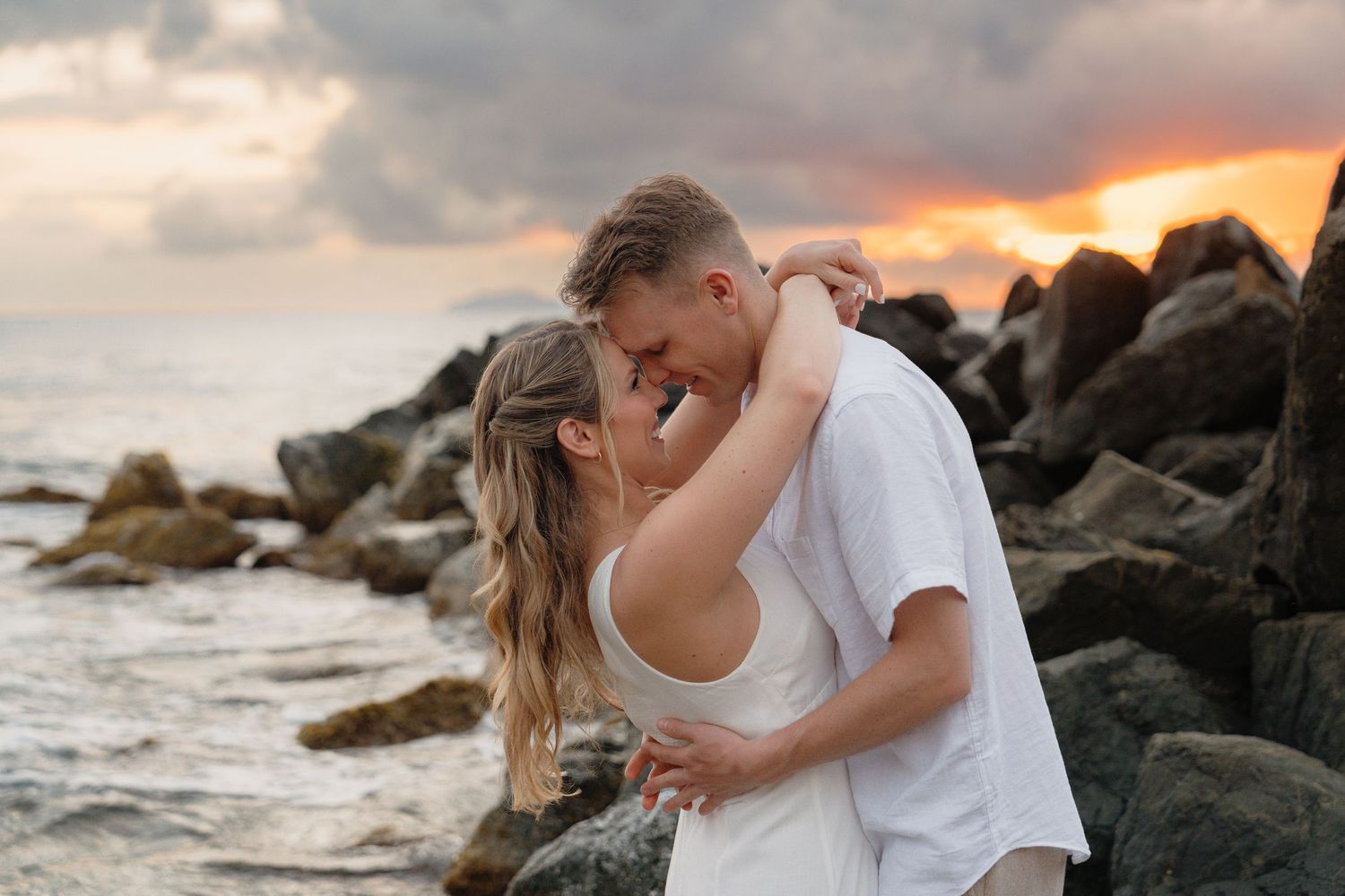 Couple sharing a romantic kiss on a rocky beach at sunset with dramatic cloudy sky in the background.
