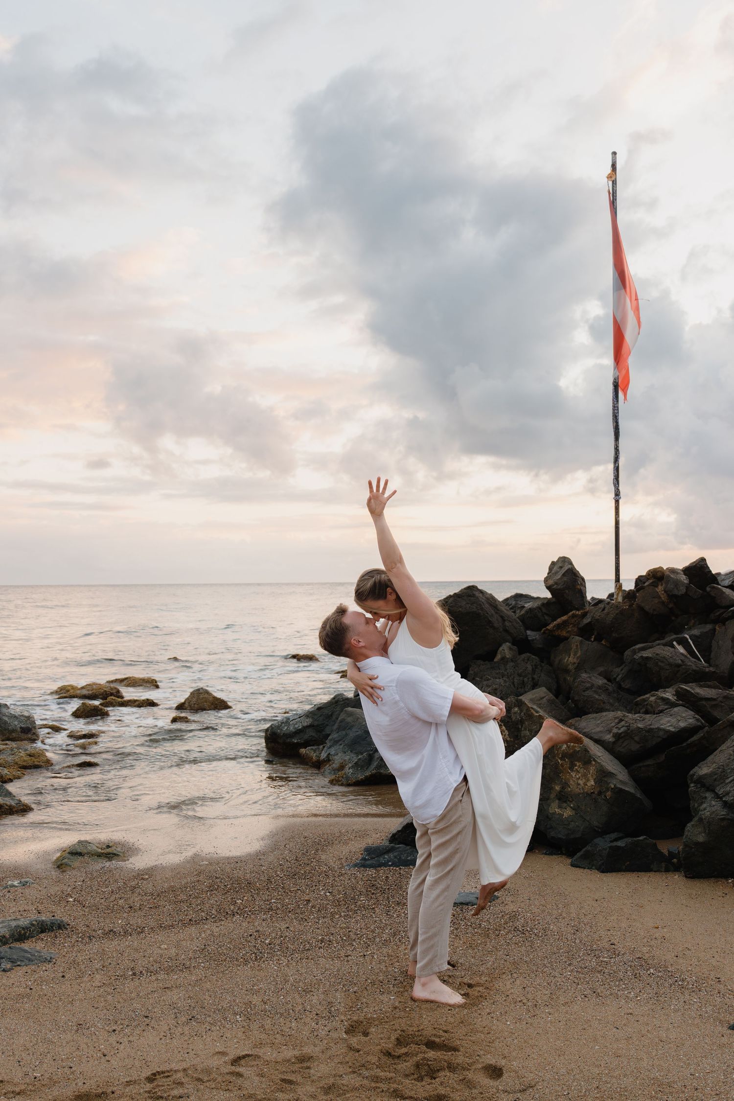 Couple dancing romantically on a sandy beach at sunset near rocky shoreline with a flag pole in the background.