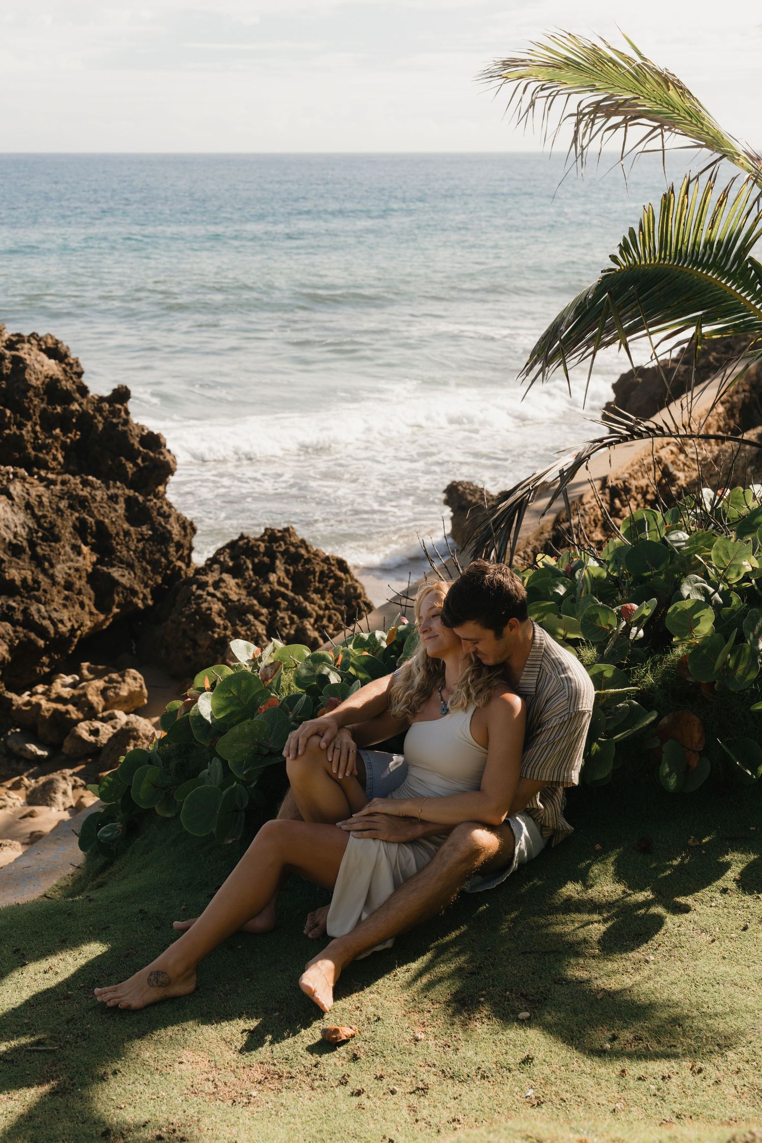 Couple sitting together on a grassy cliff by the ocean, surrounded by tropical plants and rocky shore.
