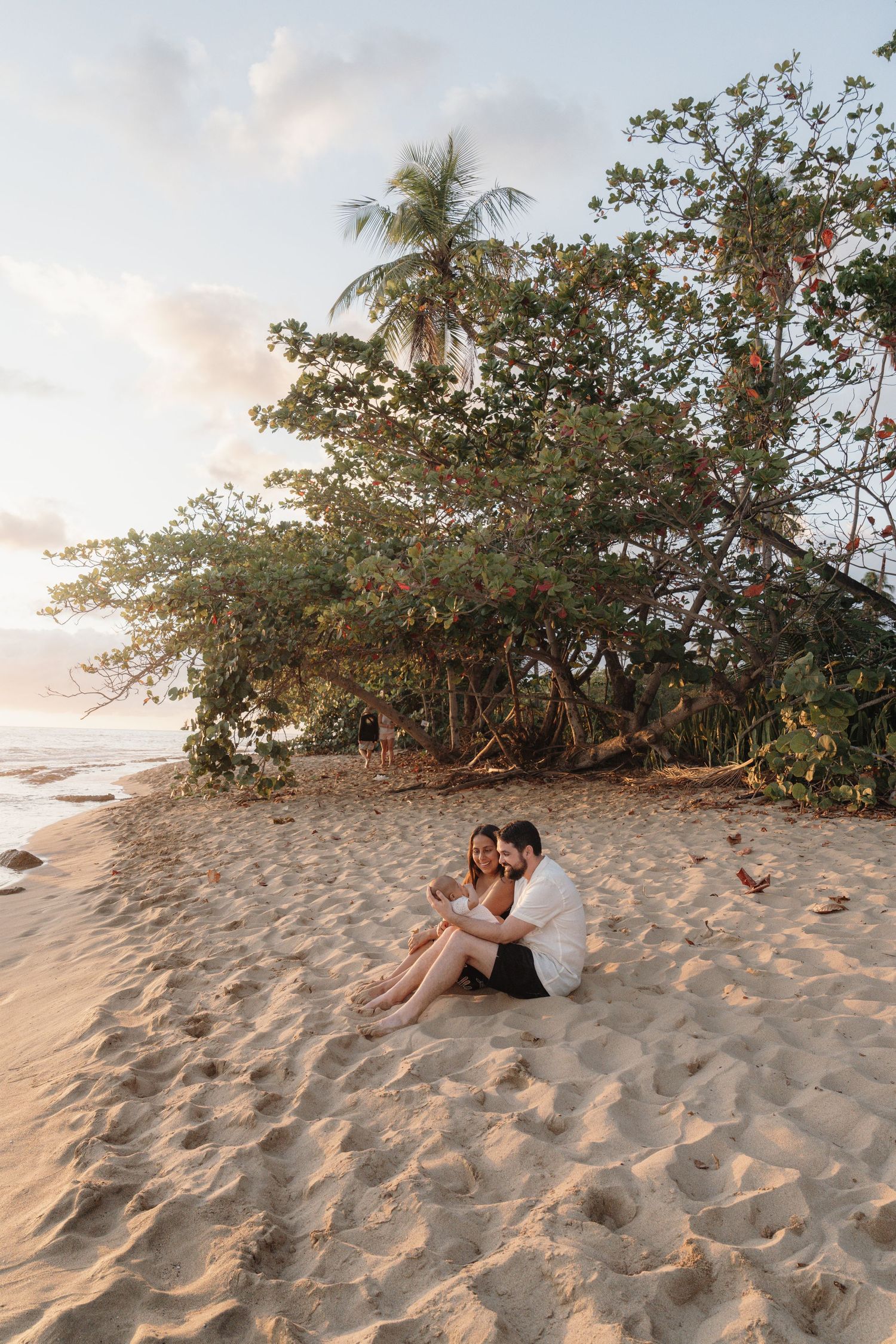Couple sitting together on a sandy tropical beach at sunset, surrounded by lush palm trees and ocean views.