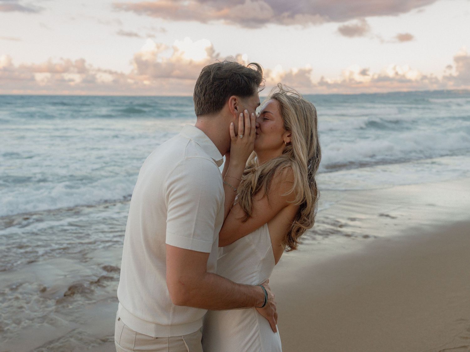 Couple sharing a romantic kiss on a tropical beach at sunset with ocean waves in the background.