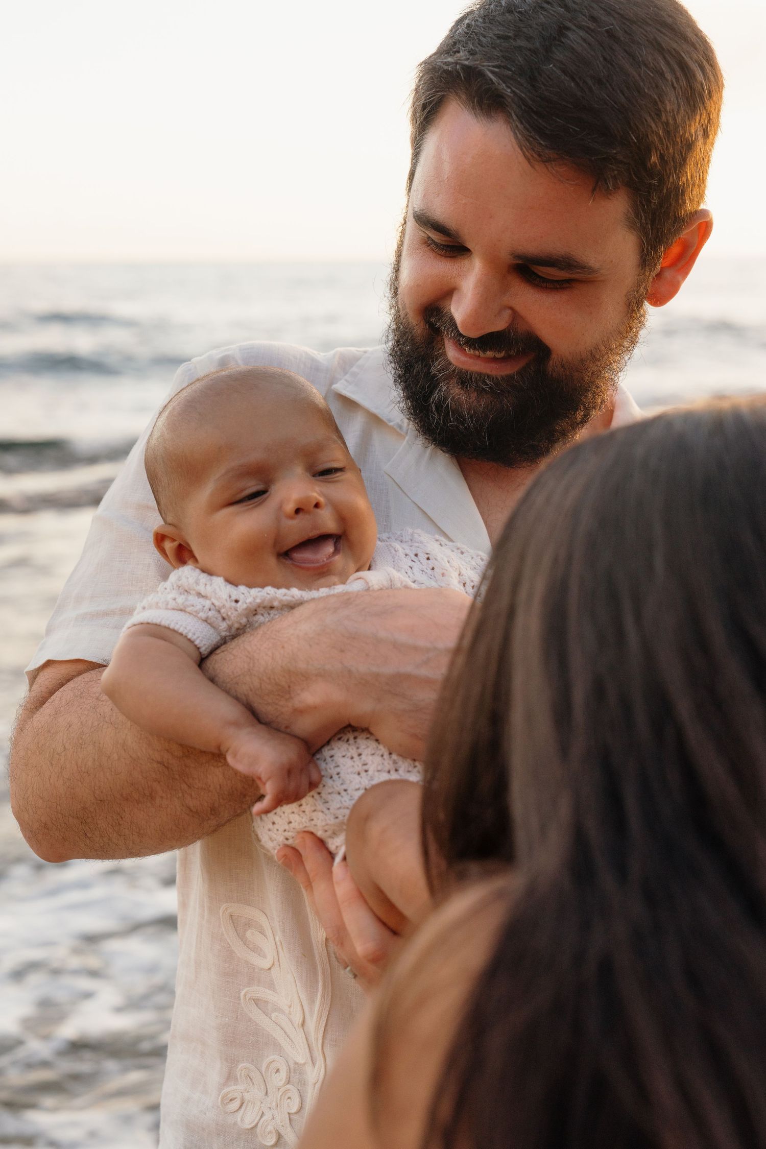 Bearded father smiling while holding laughing baby at beach sunset, mother visible in foreground.