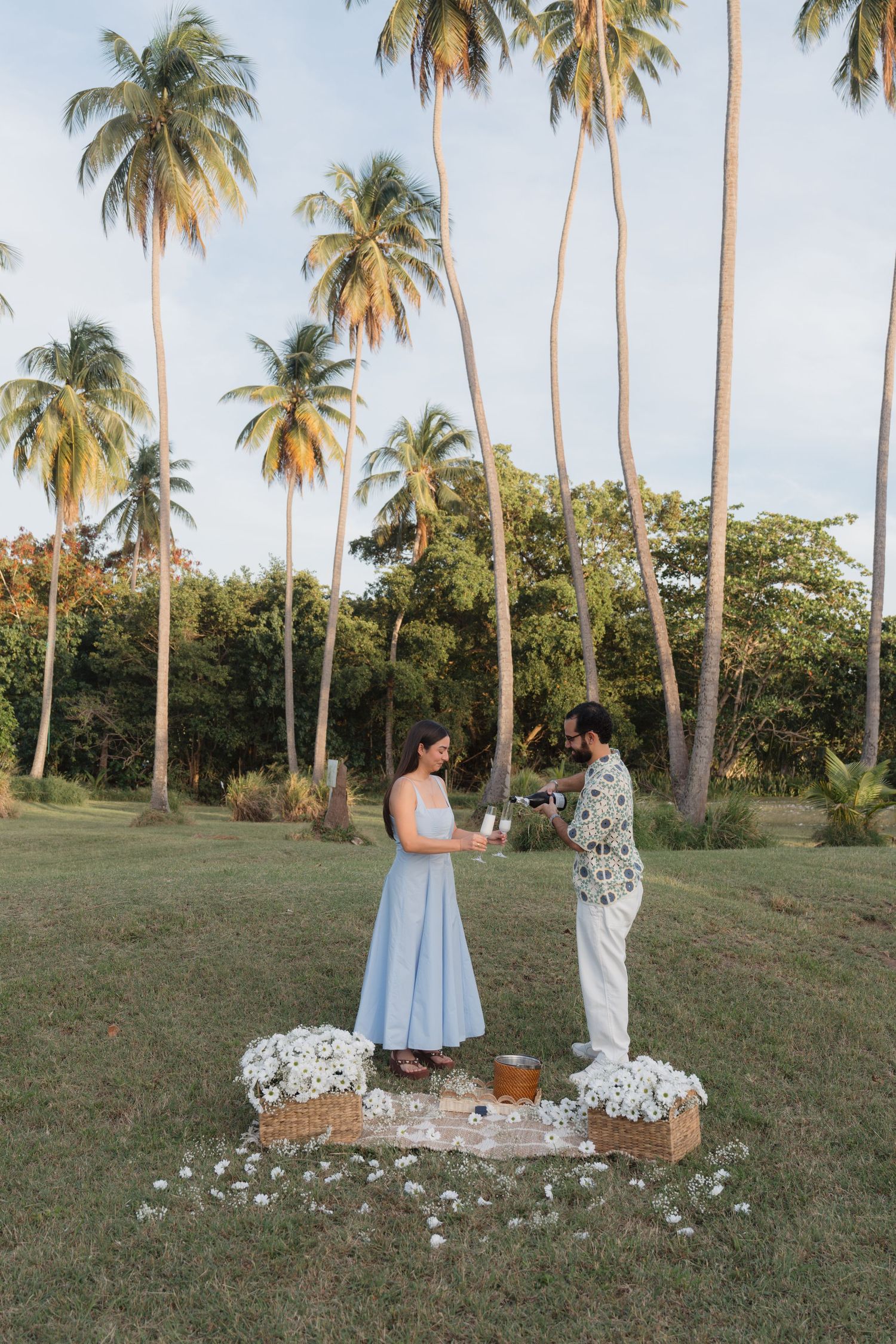 Couple sharing a romantic outdoor proposal moment surrounded by white flowers and tall palm trees at dusk.