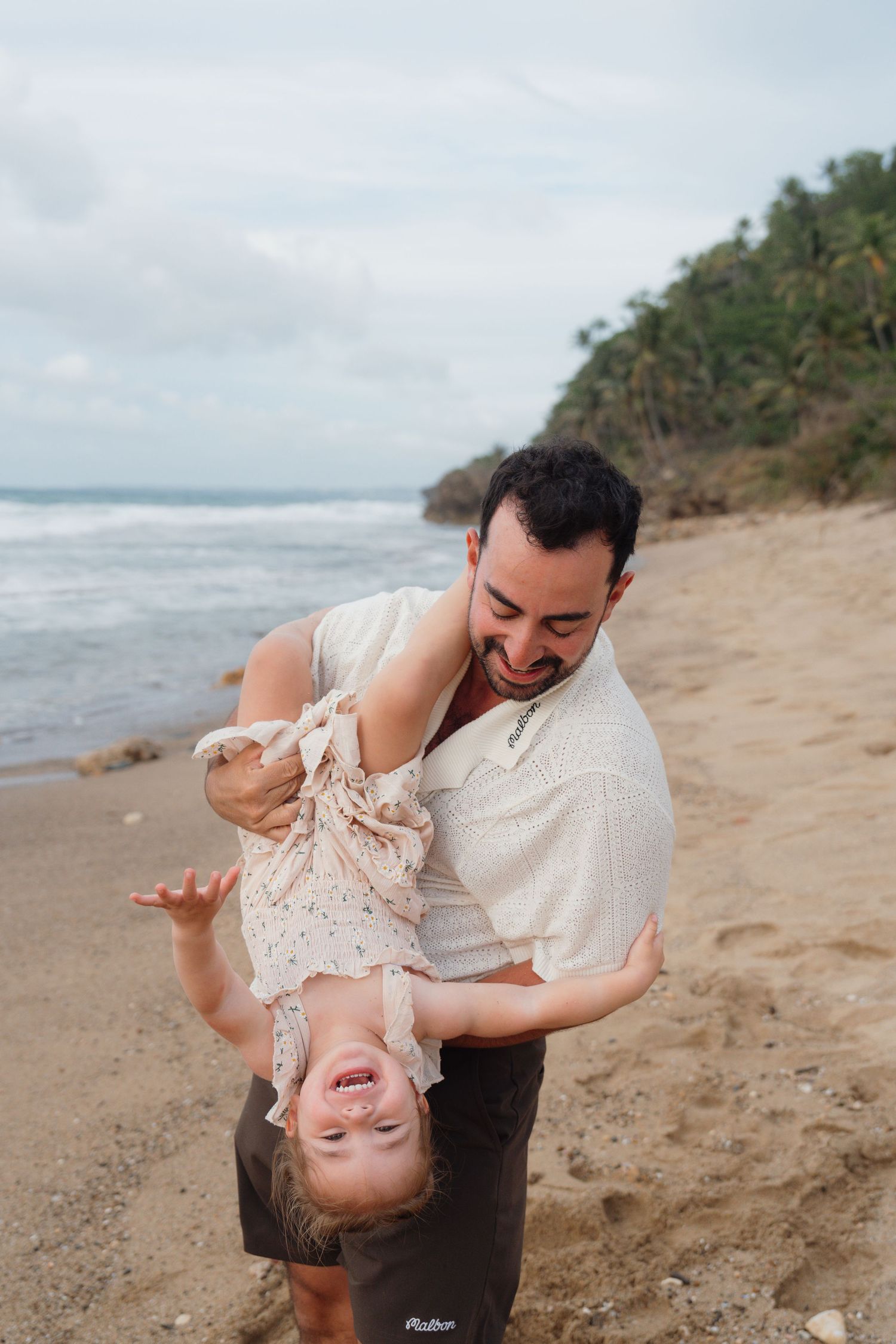 Father playfully holding toddler upside down on a tropical sandy beach, both laughing joyfully.