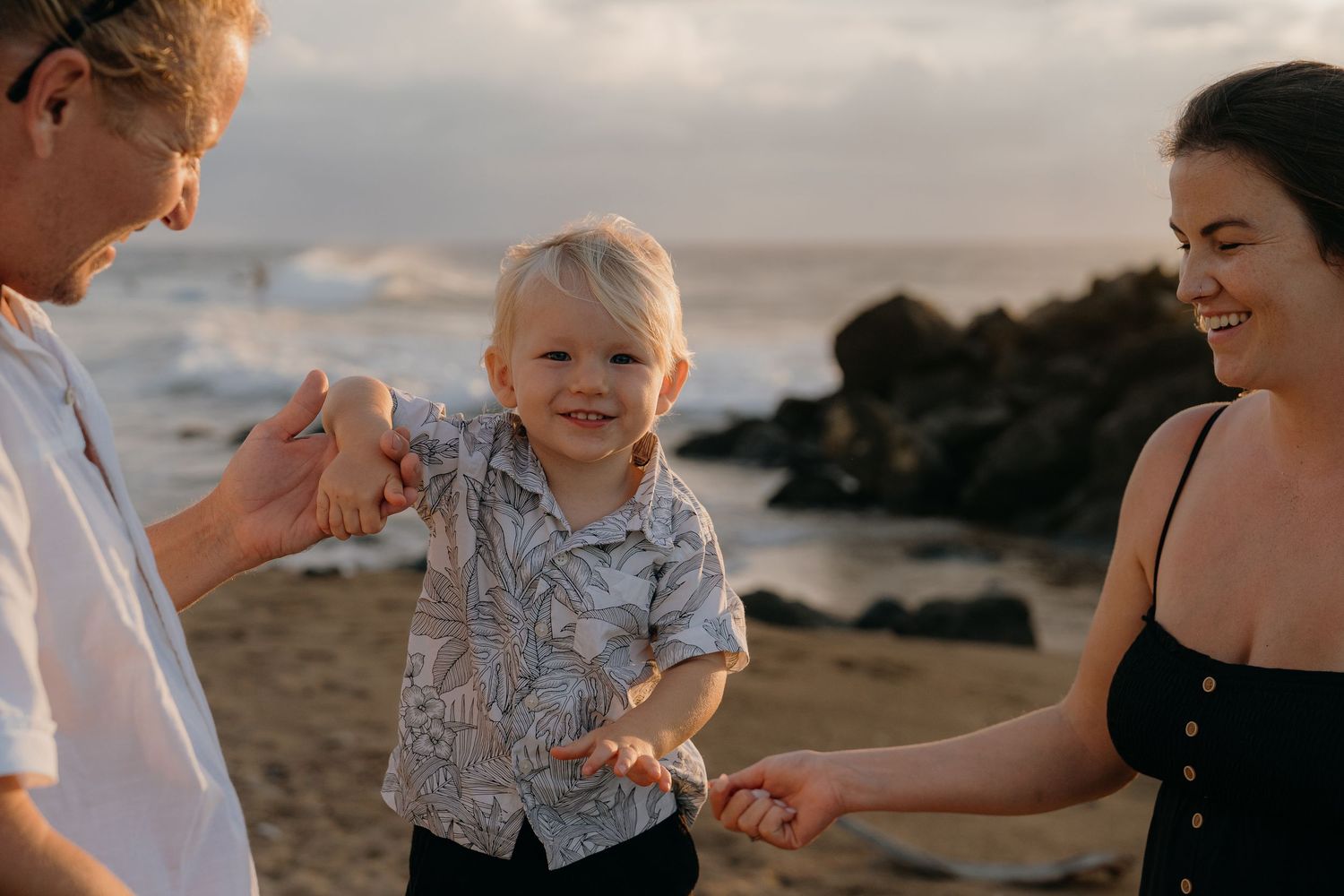 A happy toddler smiles between parents on a beach at sunset, holding hands in a warm golden light.