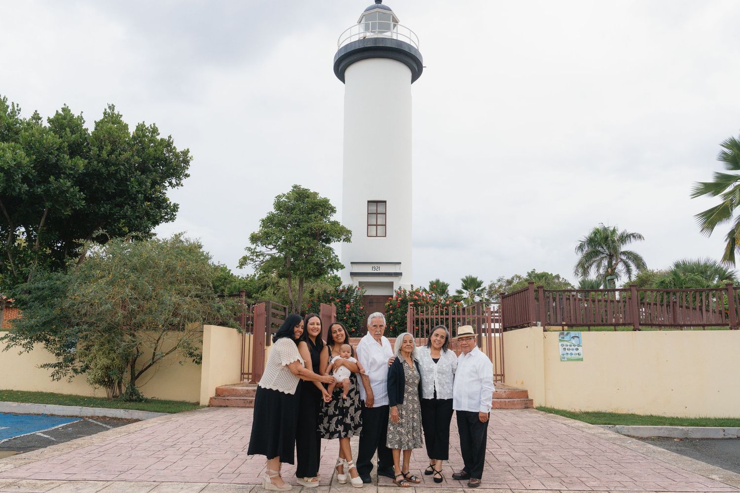Group of people posing in front of a white lighthouse surrounded by tropical trees on a sunny day.