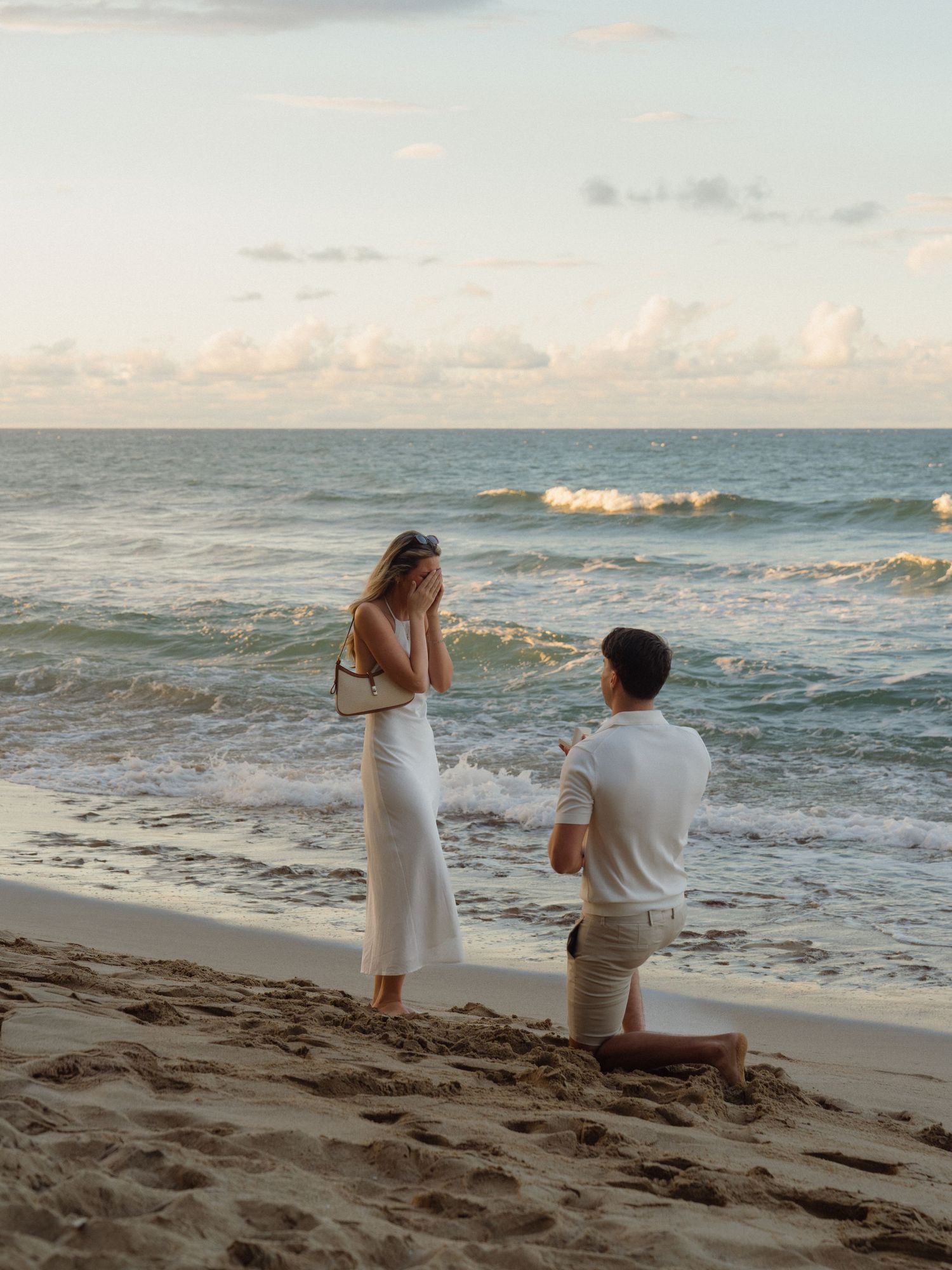 Man proposes on bended knee to woman in white dress on sandy beach at sunset with ocean waves behind them.