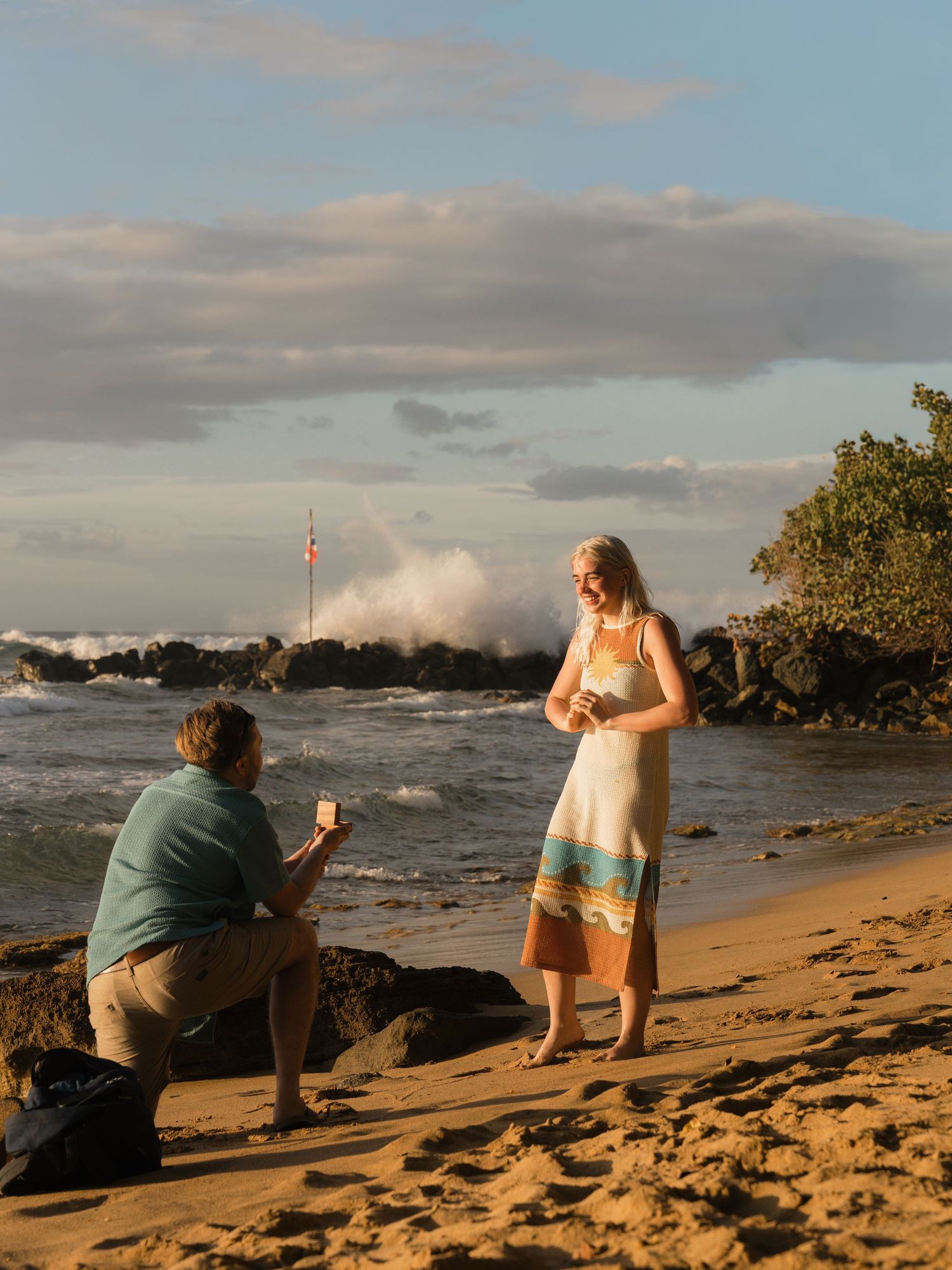 Man kneeling on sandy beach proposing to woman in striped dress at sunset with dramatic ocean waves crashing behind them.