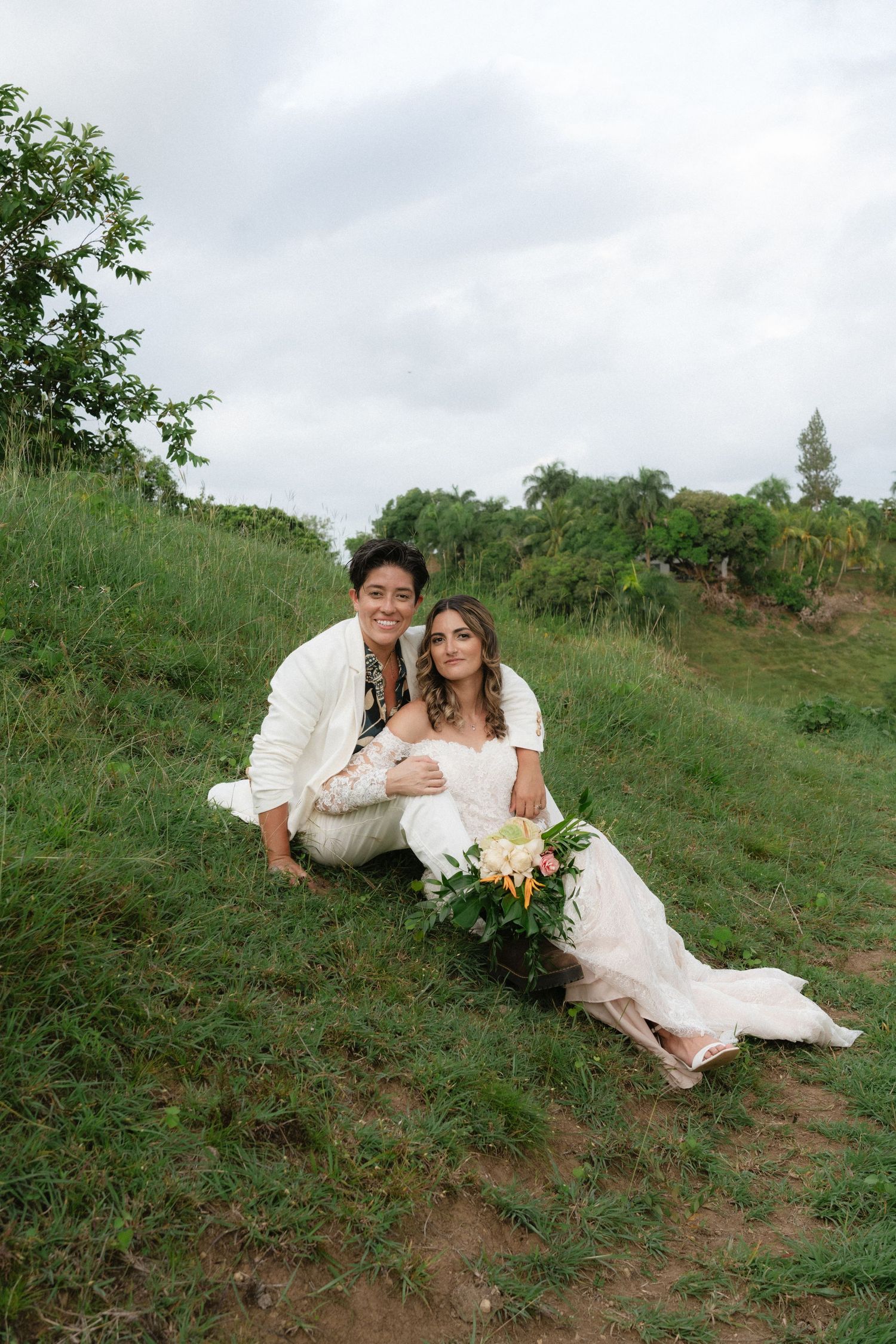 Wedding couple sitting on grassy hillside in elegant attire.