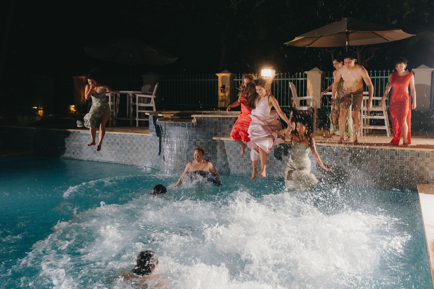 People jumping and splashing in an illuminated outdoor pool at night during a lively party gathering.