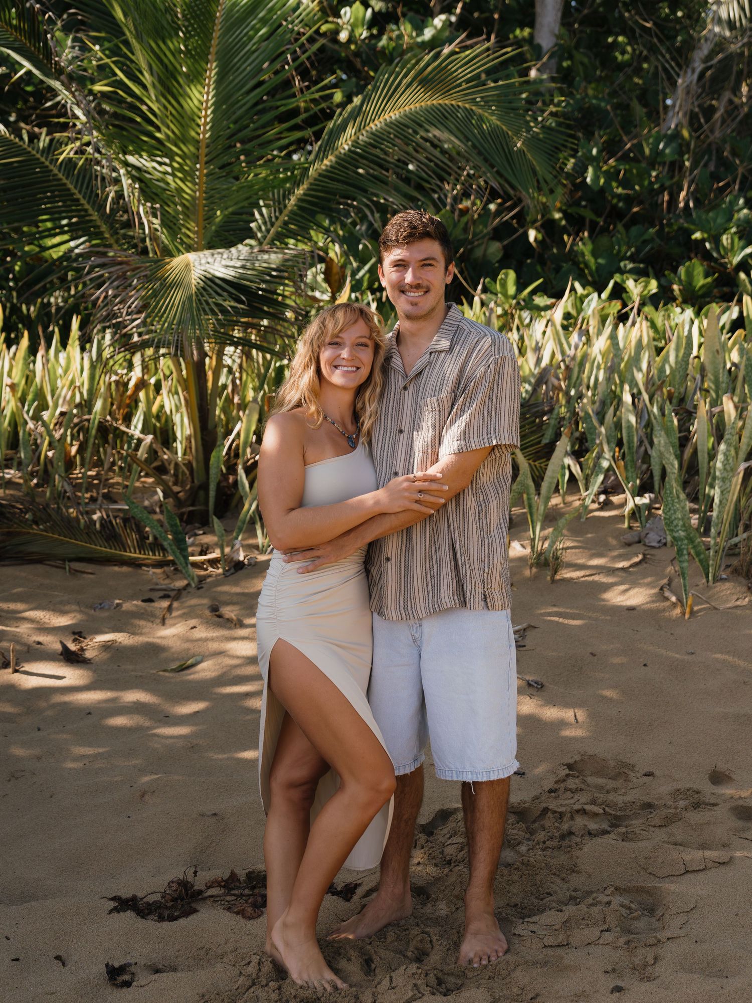 Couple embracing on a tropical beach surrounded by lush palm trees and green foliage on a sunny day.