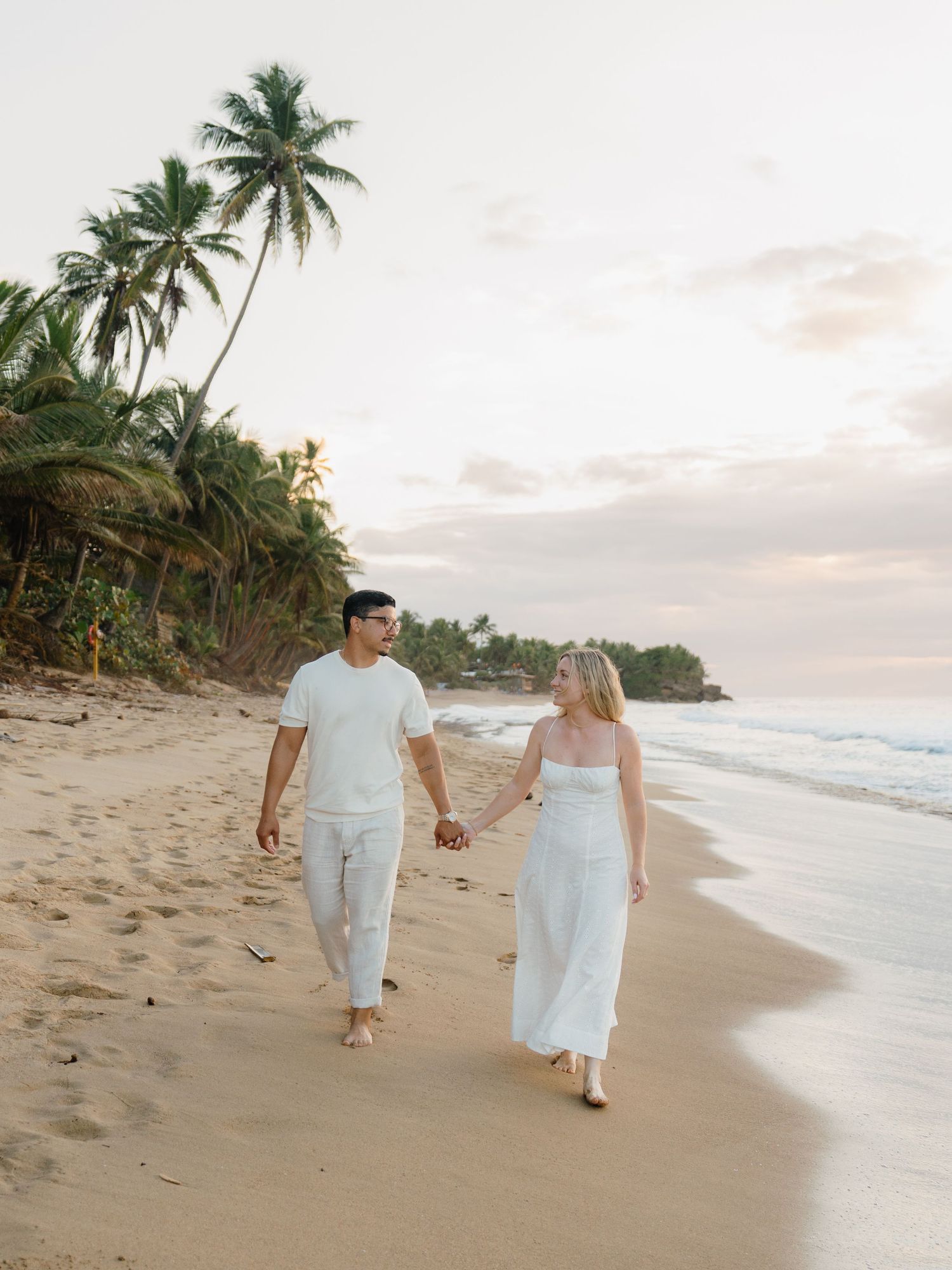 Couple in white outfits holding hands walking on a tropical beach with palm trees at sunset.