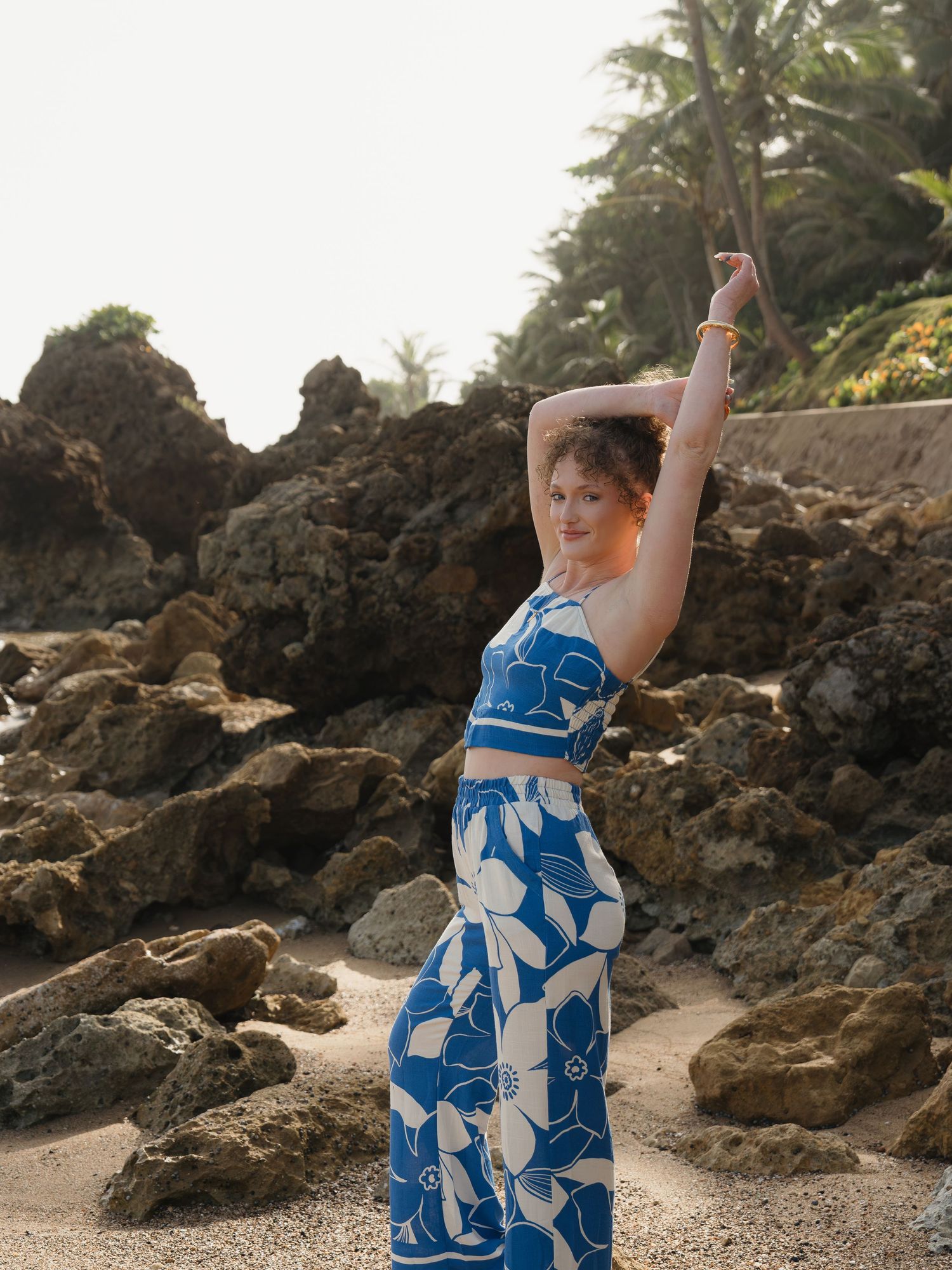 Woman in blue floral crop top and wide-leg pants posing on rocky beach with palm trees.