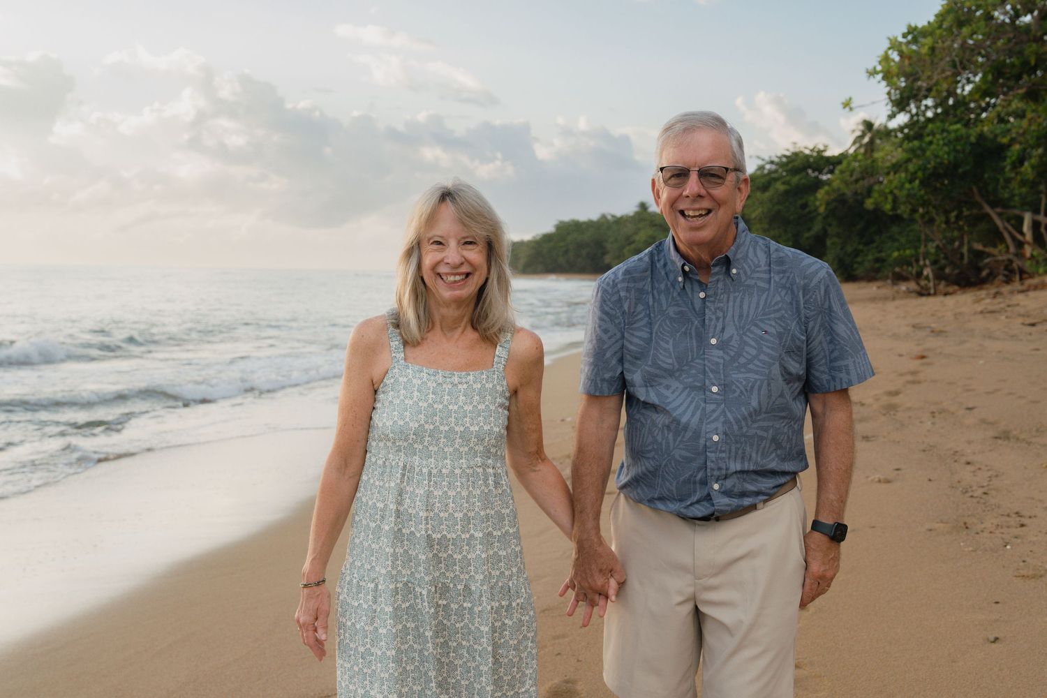 Smiling senior couple holding hands on a tropical beach at sunset with waves and lush trees in the background.