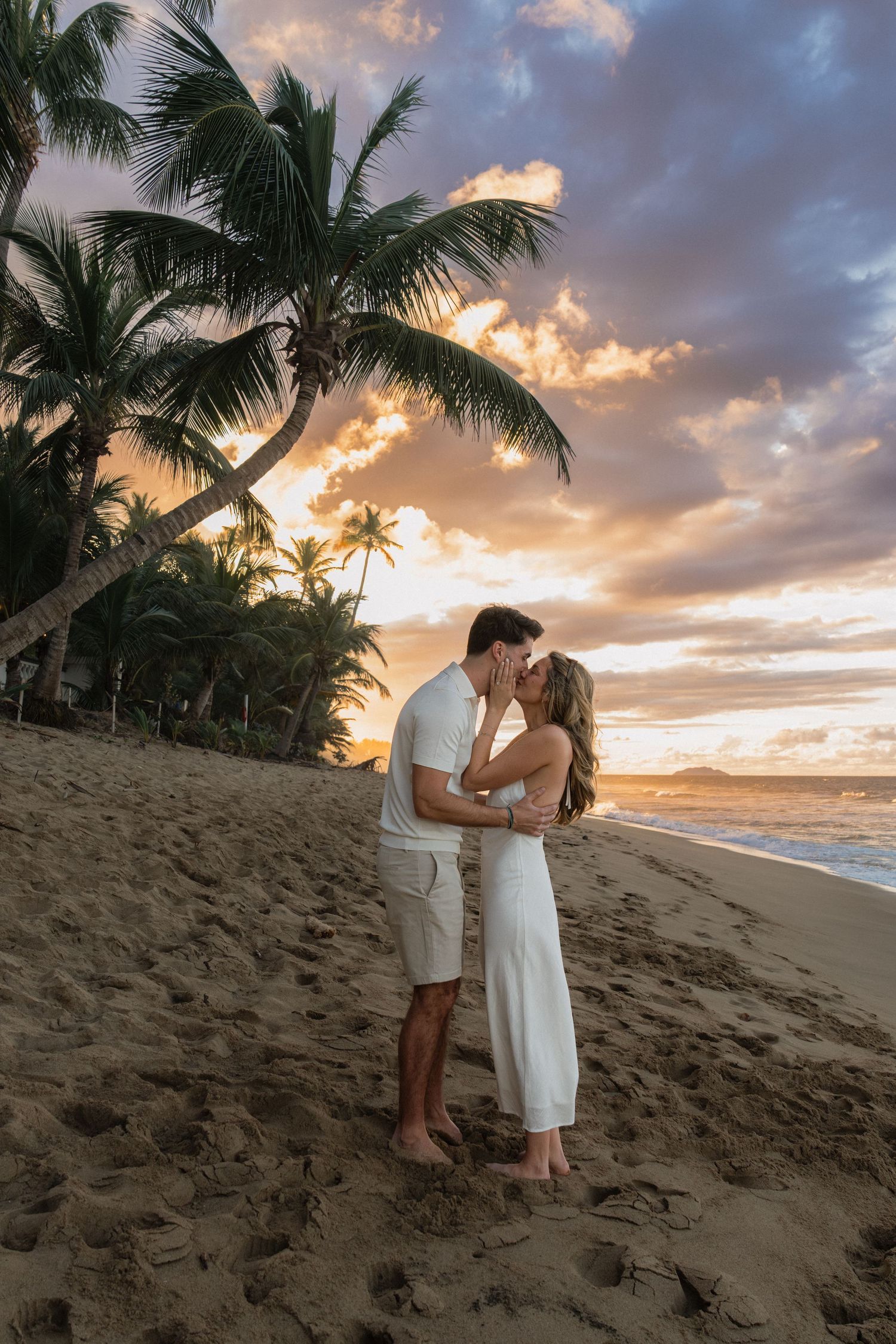 Couple kissing on a tropical beach at sunset with palm trees and colorful sky in the background.