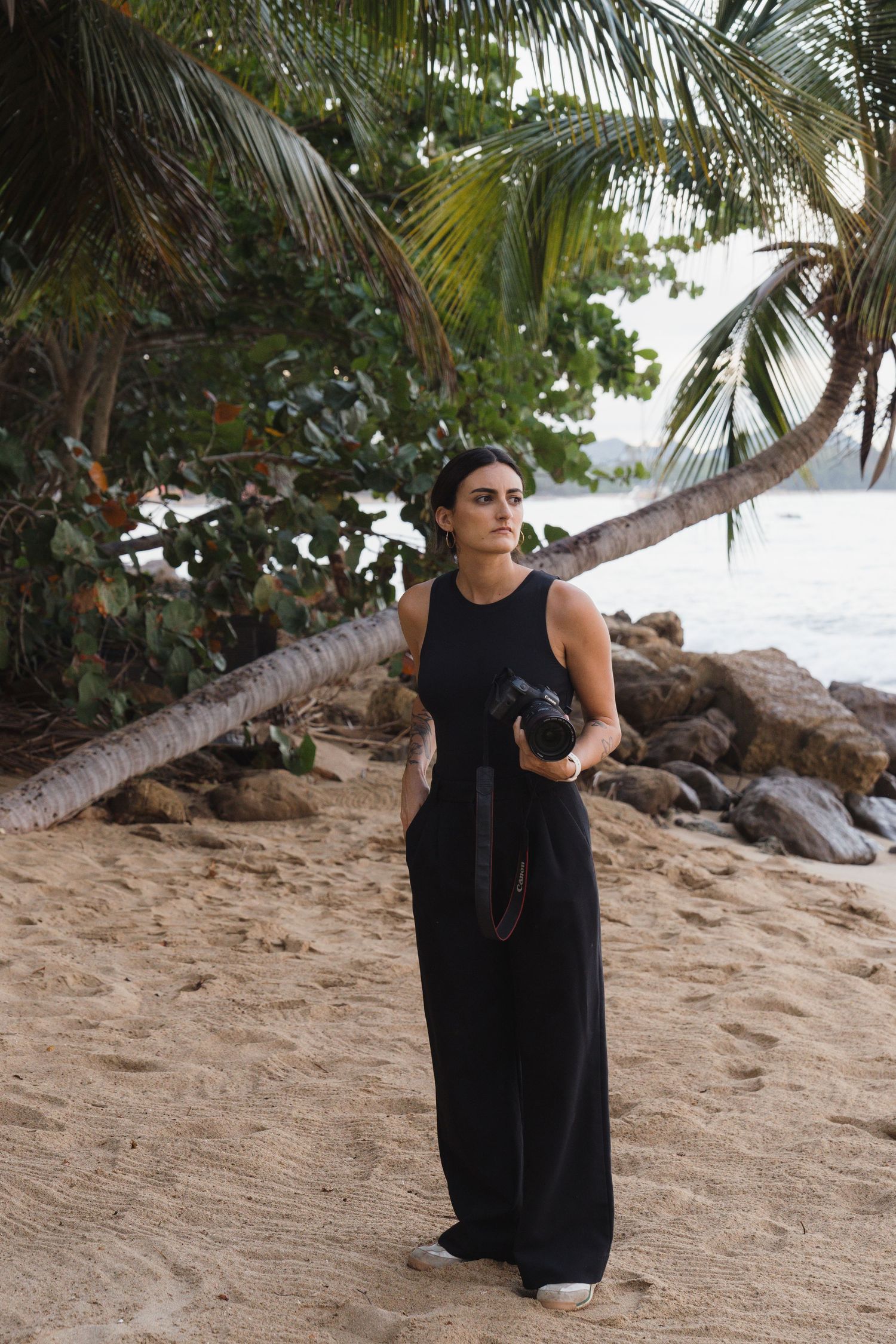 Woman in black jumpsuit holding a camera on a tropical beach with palm trees and rocky shoreline.