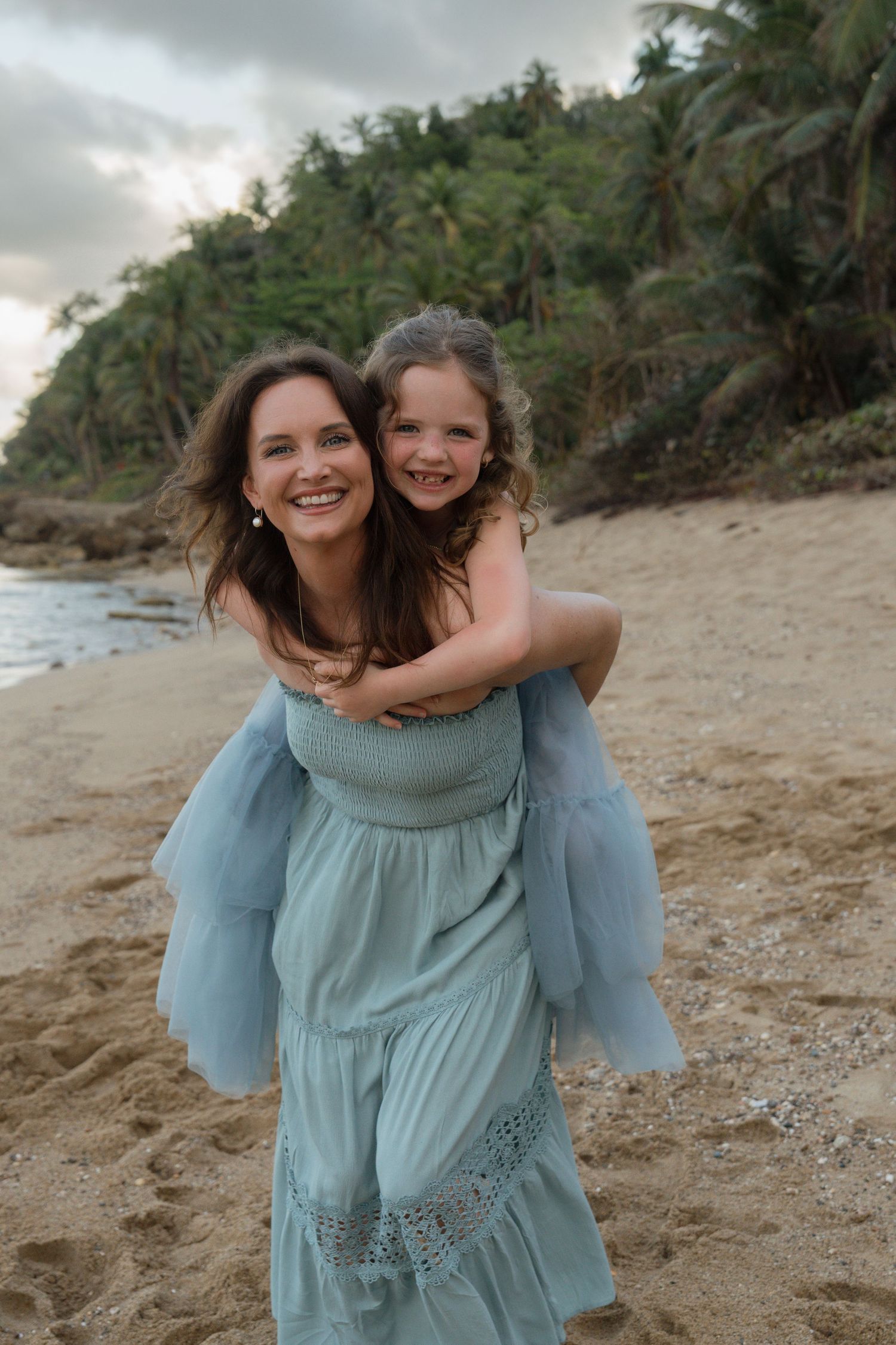 A woman in a light blue dress gives a young girl a piggyback ride on a tropical beach with lush green palms.